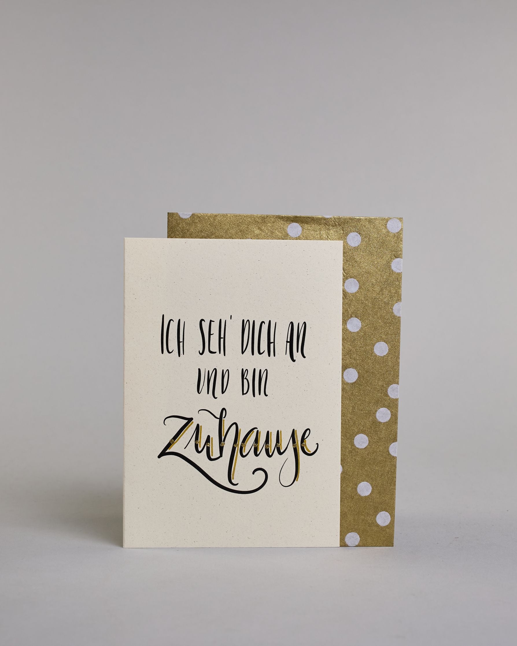 Klappkarte für Liebende mit schwarzer Kalligrafieaufschrift "Ich seh' dich an und bin Zuhause" auf hellem Hintergrund. Der Text ist in folgende drei Zeilen aufgeteilt: "ICH SEH' DICH AN", darunter "UND BIN" beide Zeilen sind in einer verspielten geraden Kalligrafieschrift gestaltet, darunter "Zuhause" in verspielter geschwungener Schreibschrift. Dazu ein handgeschöpftes Kuvert. #2