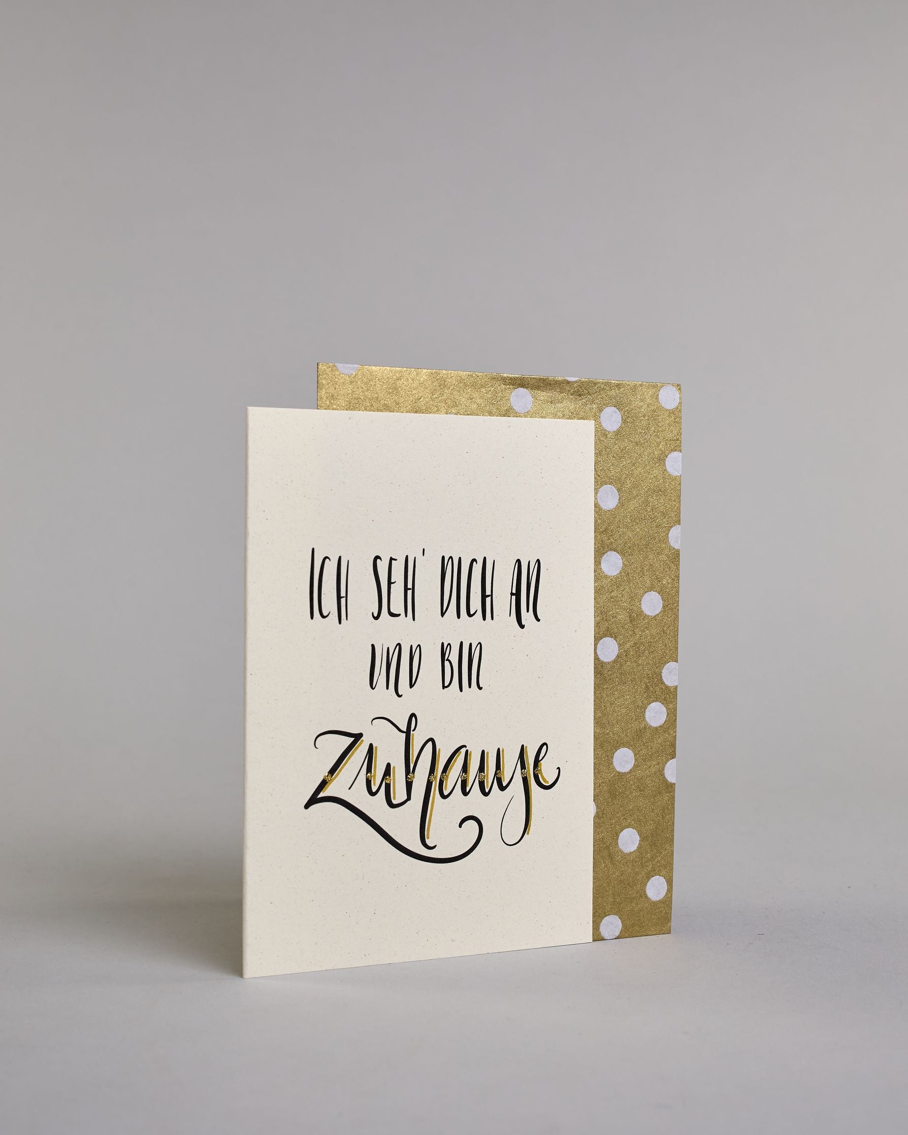 Klappkarte für Liebende mit schwarzer Kalligrafieaufschrift "Ich seh' dich an und bin Zuhause" auf hellem Hintergrund. Der Text ist in folgende drei Zeilen aufgeteilt: "ICH SEH' DICH AN", darunter "UND BIN" beide Zeilen sind in einer verspielten geraden Kalligrafieschrift gestaltet, darunter "Zuhause" in verspielter geschwungener Schreibschrift. Dazu ein handgeschöpftes Kuvert. #3