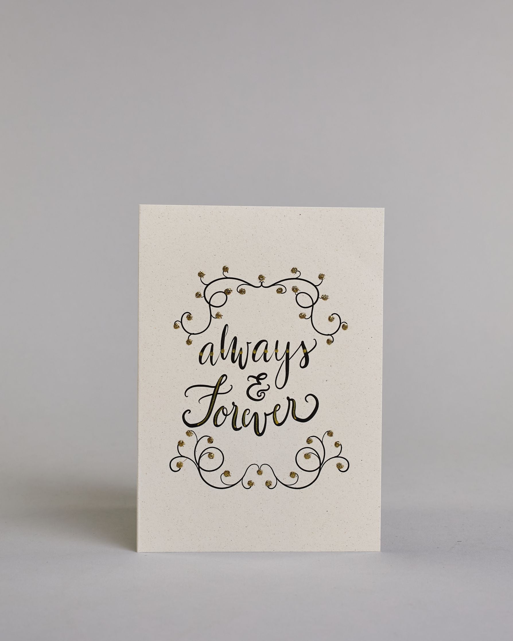 Klappkarte für Liebende in Englisch mit schwarzer Kalligrafieaufschrift "Always & Forever" mit verschnörkelten Verzierungen über und unter dem Text auf hellem Hintergrund. Der Text ist in folgende drei Zeilen aufgeteilt: "always", darunter "&", darunter "forever" alles ist in einer verschnörkelten Schreibschrift mit punktueller goldener und Glitzerverzierung gestaltet. Dazu ein gold-weißes handgeschöpftes Kuvert. #1