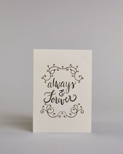 Klappkarte für Liebende in Englisch mit schwarzer Kalligrafieaufschrift "Always & Forever" mit verschnörkelten Verzierungen über und unter dem Text auf hellem Hintergrund. Der Text ist in folgende drei Zeilen aufgeteilt: "always", darunter "&", darunter "forever" alles ist in einer verschnörkelten Schreibschrift mit punktueller goldener und Glitzerverzierung gestaltet. Dazu ein gold-weißes handgeschöpftes Kuvert. #1