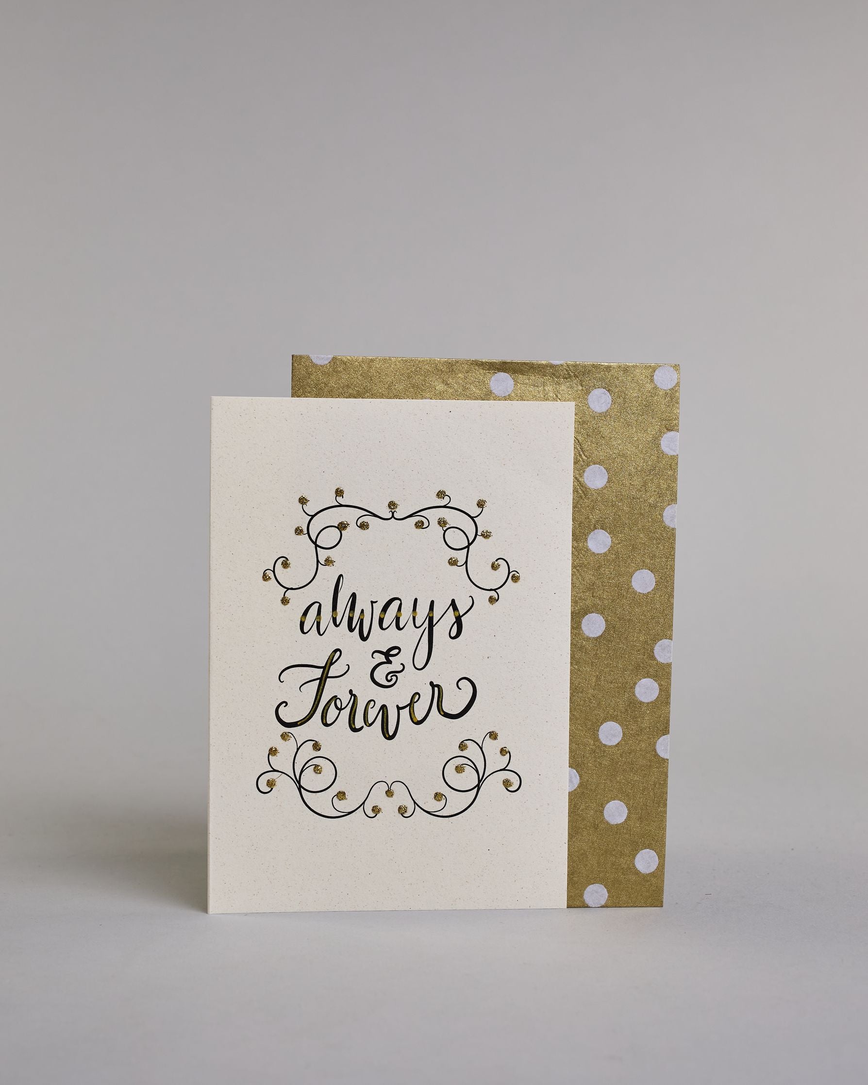 Klappkarte für Liebende in Englisch mit schwarzer Kalligrafieaufschrift "Always & Forever" mit verschnörkelten Verzierungen über und unter dem Text auf hellem Hintergrund. Der Text ist in folgende drei Zeilen aufgeteilt: "always", darunter "&", darunter "forever" alles ist in einer verschnörkelten Schreibschrift mit punktueller goldener und Glitzerverzierung gestaltet. Dazu ein gold-weißes handgeschöpftes Kuvert. #2