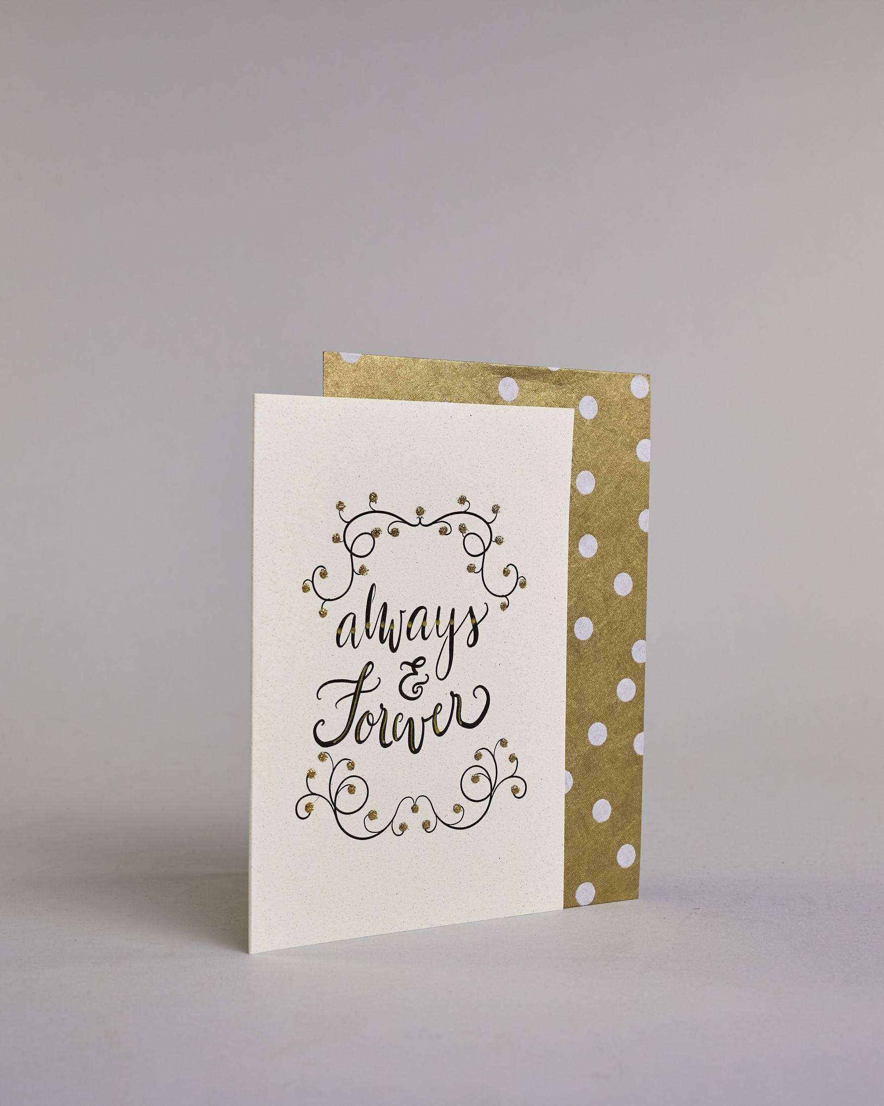 Klappkarte für Liebende in Englisch mit schwarzer Kalligrafieaufschrift "Always & Forever" mit verschnörkelten Verzierungen über und unter dem Text auf hellem Hintergrund. Der Text ist in folgende drei Zeilen aufgeteilt: "always", darunter "&", darunter "forever" alles ist in einer verschnörkelten Schreibschrift mit punktueller goldener und Glitzerverzierung gestaltet. Dazu ein gold-weißes handgeschöpftes Kuvert. #3