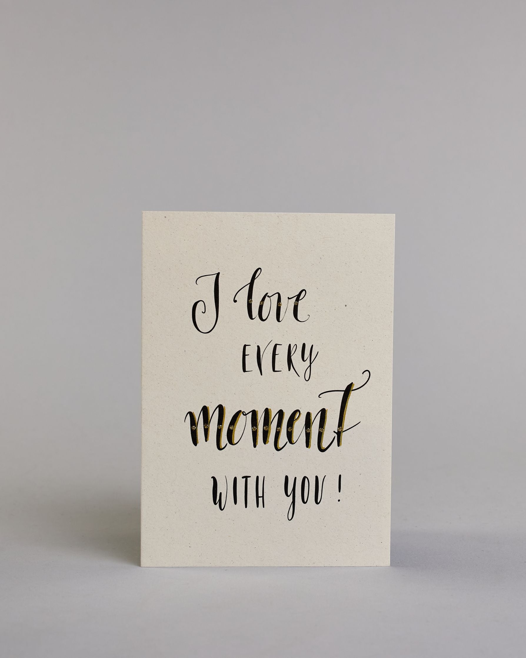 Klappkarte für Liebende in Englisch mit schwarzer Kalligrafieaufschrift "I love every moment with you!" auf hellem Hintergrund. Der Text ist in folgende vier Zeilen aufgeteilt: "I love" in verschnörkelter Schreibschrift mit glitzernden Punkten auf dem Wort "love", darunter "EVERY" in verspielter gerader Kalligrafieschrift, darunter "moment" größer gehalten in einer dickeren Schreibschrift, darunter "with you!" in der gleichen Schrift wie das Wort "EVERY". Dazu ein grün-goldenes handgeschöpftes Kuvert. #1