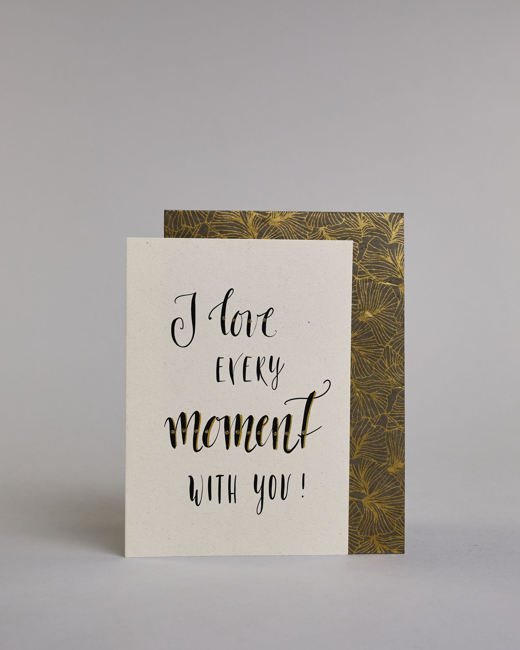 Klappkarte für Liebende in Englisch mit schwarzer Kalligrafieaufschrift "I love every moment with you!" auf hellem Hintergrund. Der Text ist in folgende vier Zeilen aufgeteilt: "I love" in verschnörkelter Schreibschrift mit glitzernden Punkten auf dem Wort "love", darunter "EVERY" in verspielter gerader Kalligrafieschrift, darunter "moment" größer gehalten in einer dickeren Schreibschrift, darunter "with you!" in der gleichen Schrift wie das Wort "EVERY". Dazu ein grün-goldenes handgeschöpftes Kuvert. #2