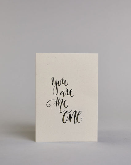 Klappkarte für Liebende in Englisch mit schwarzer Kalligrafieaufschrift "You are the one" auf hellem Hintergrund. Der Text ist in vier Zeilen mit jeweils einem Wort aufgeteilt und in einer geschwungenen Schreibschrift mit Glitzerverzierung gestaltet. Dazu ein gold-weißes handgeschöpftes Kuvert. #1