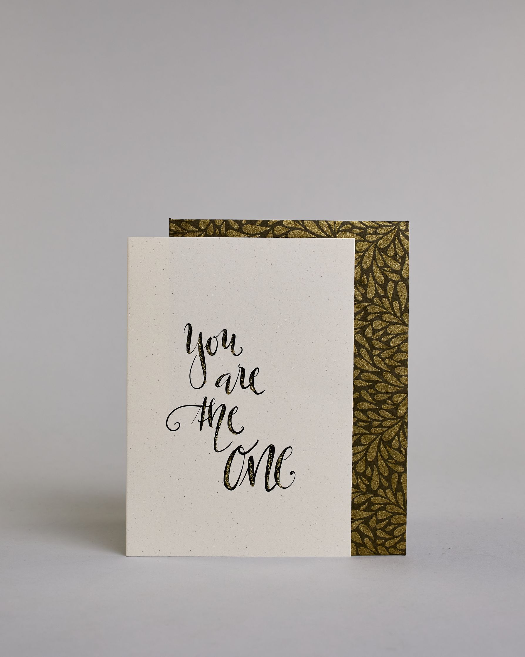 Klappkarte für Liebende in Englisch mit schwarzer Kalligrafieaufschrift "You are the one" auf hellem Hintergrund. Der Text ist in vier Zeilen mit jeweils einem Wort aufgeteilt und in einer geschwungenen Schreibschrift mit Glitzerverzierung gestaltet. Dazu ein gold-weißes handgeschöpftes Kuvert. #2
