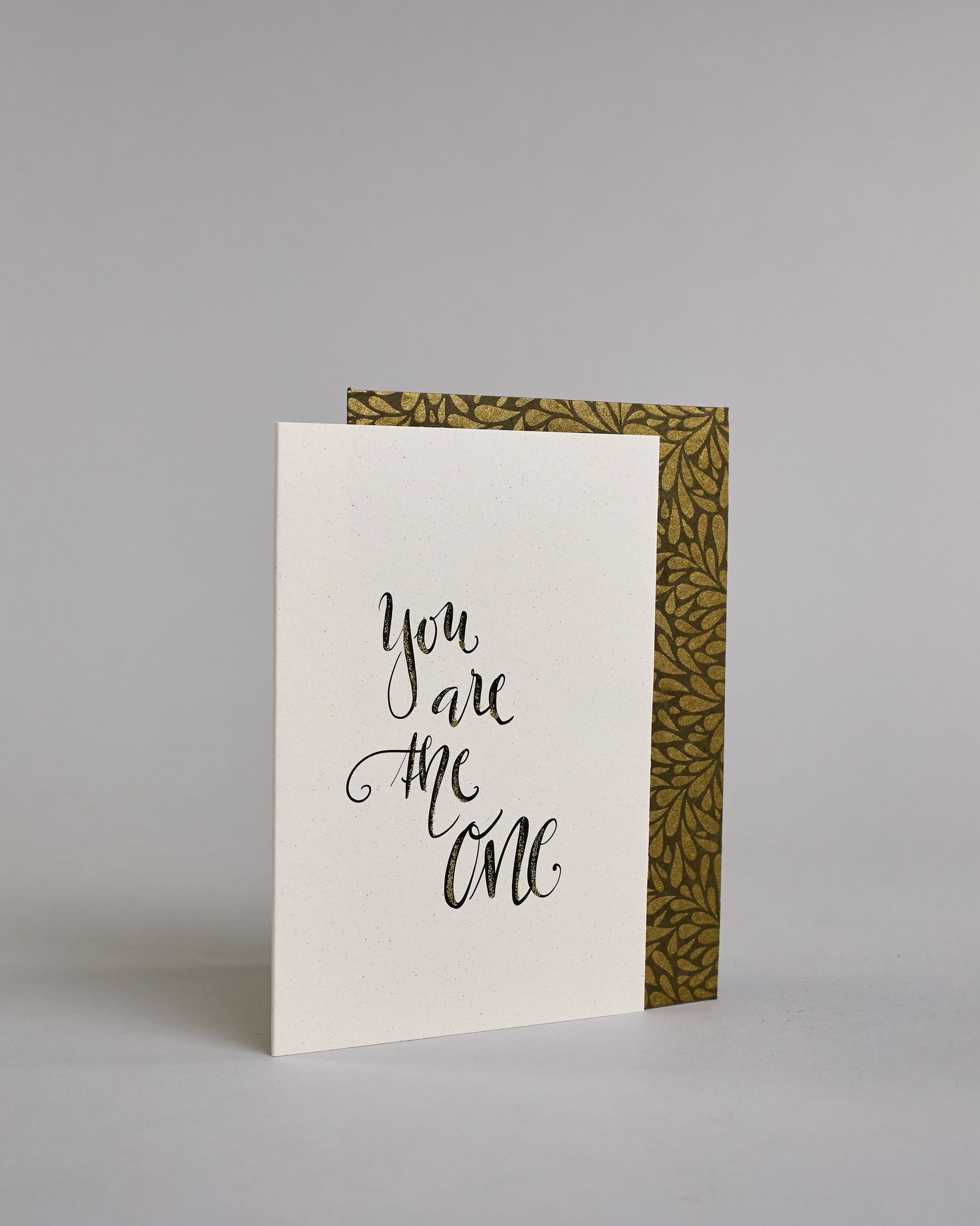 Klappkarte für Liebende in Englisch mit schwarzer Kalligrafieaufschrift "You are the one" auf hellem Hintergrund. Der Text ist in vier Zeilen mit jeweils einem Wort aufgeteilt und in einer geschwungenen Schreibschrift mit Glitzerverzierung gestaltet. Dazu ein gold-weißes handgeschöpftes Kuvert. #3