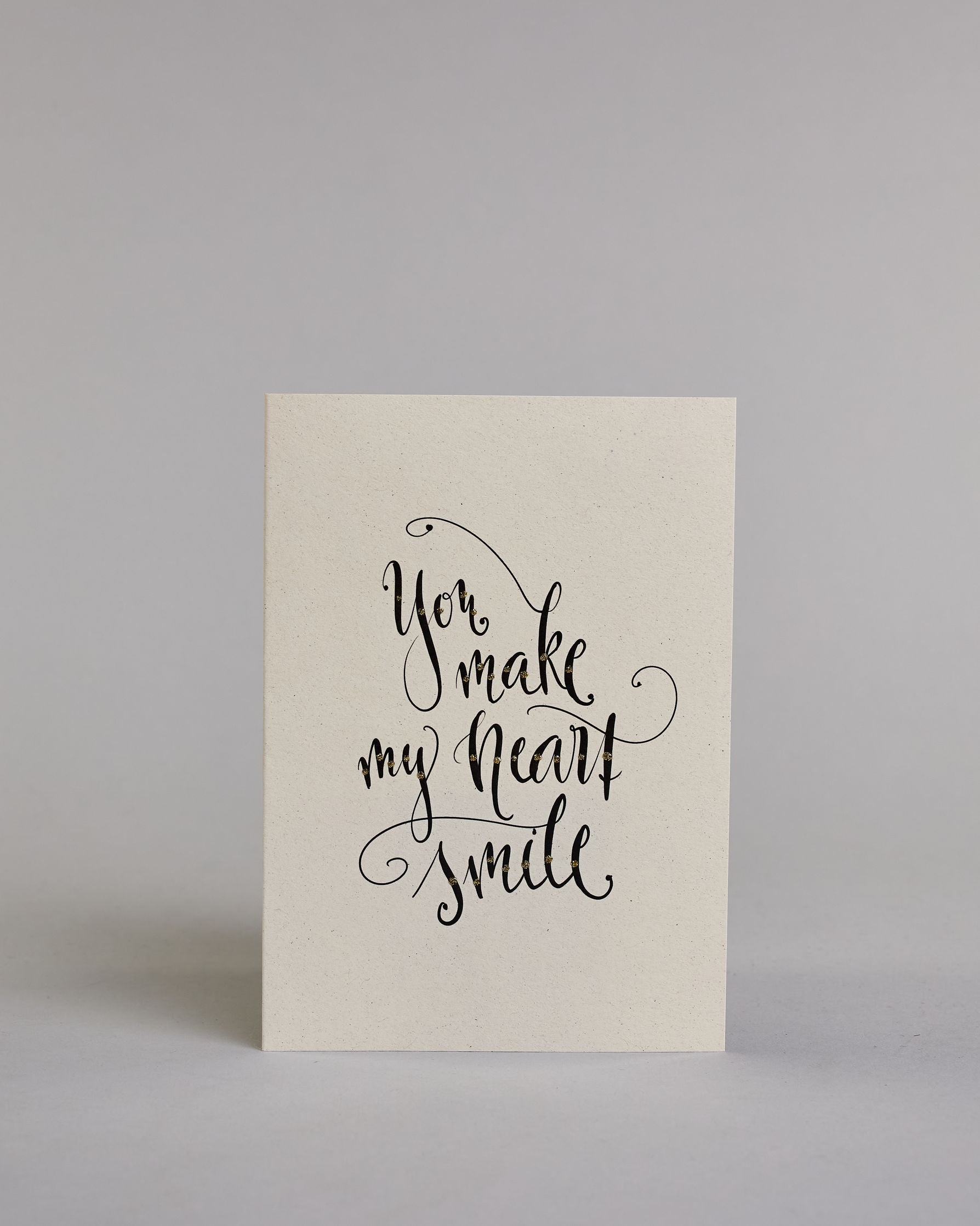 Klappkarte für Liebende in Englisch mit schwarzer Kalligrafieaufschrift "You make my heart smile" auf hellem Hintergrund. Der Text ist in folgende vier Zeilen aufgeteilt: "you", darunter "make", darunter "my heart", darunter "smile". Der Text ist in einer geschwungenen Schreibschrift mit punktueller Glitzerverzierung in der Mitte der Buchstaben gestaltet. Dazu ein grün-goldenes handgeschöpftes Kuvert. #1