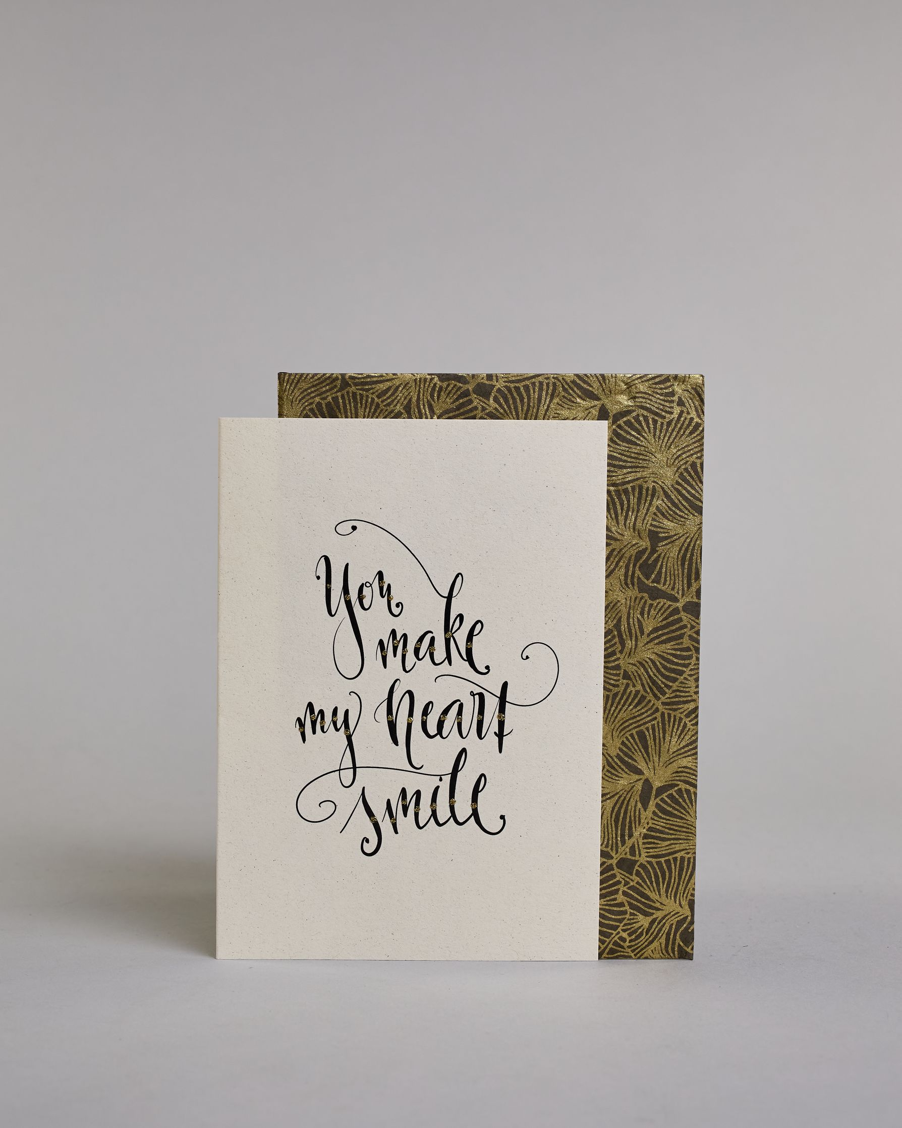 Klappkarte für Liebende in Englisch mit schwarzer Kalligrafieaufschrift "You make my heart smile" auf hellem Hintergrund. Der Text ist in folgende vier Zeilen aufgeteilt: "you", darunter "make", darunter "my heart", darunter "smile". Der Text ist in einer geschwungenen Schreibschrift mit punktueller Glitzerverzierung in der Mitte der Buchstaben gestaltet. Dazu ein grün-goldenes handgeschöpftes Kuvert. #2