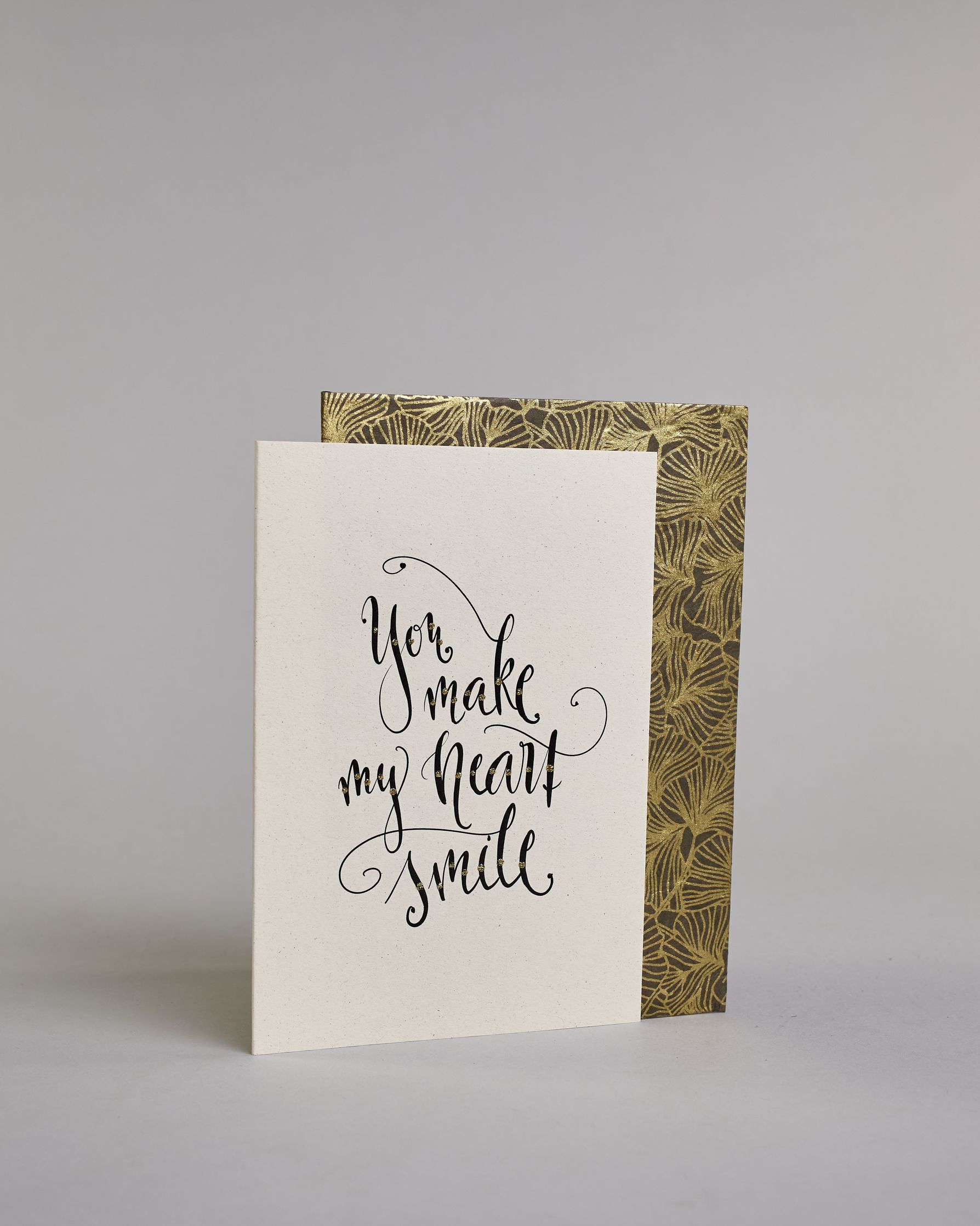 Klappkarte für Liebende in Englisch mit schwarzer Kalligrafieaufschrift "You make my heart smile" auf hellem Hintergrund. Der Text ist in folgende vier Zeilen aufgeteilt: "you", darunter "make", darunter "my heart", darunter "smile". Der Text ist in einer geschwungenen Schreibschrift mit punktueller Glitzerverzierung in der Mitte der Buchstaben gestaltet. Dazu ein grün-goldenes handgeschöpftes Kuvert. #3