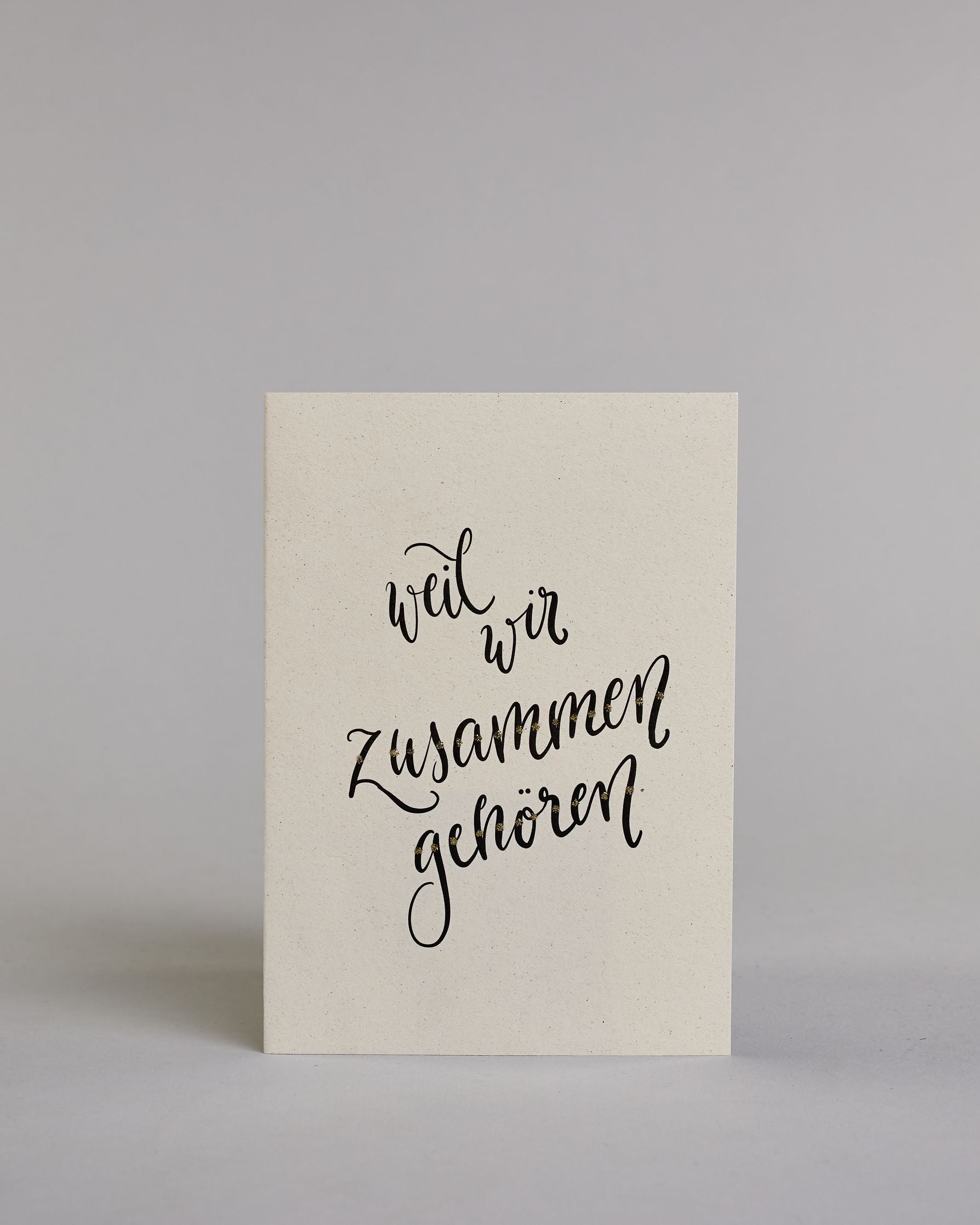 Klappkarte für Liebende mit schwarzer Kalligrafieaufschrift "weil wir zusammen gehören" auf hellem Hintergrund. Der Text ist in vier Zeilen mit jeweils einem Worrt aufgeteilt. Der Text ist in einer geschwungenen Schreibschrift mit punktueller Glitzerverzierung in der Mitte der Buchstaben gestaltet. Dazu ein grün-goldenes handgeschöpftes Kuvert. #1