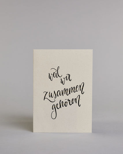 Klappkarte für Liebende mit schwarzer Kalligrafieaufschrift "weil wir zusammen gehören" auf hellem Hintergrund. Der Text ist in vier Zeilen mit jeweils einem Worrt aufgeteilt. Der Text ist in einer geschwungenen Schreibschrift mit punktueller Glitzerverzierung in der Mitte der Buchstaben gestaltet. Dazu ein grün-goldenes handgeschöpftes Kuvert. #1