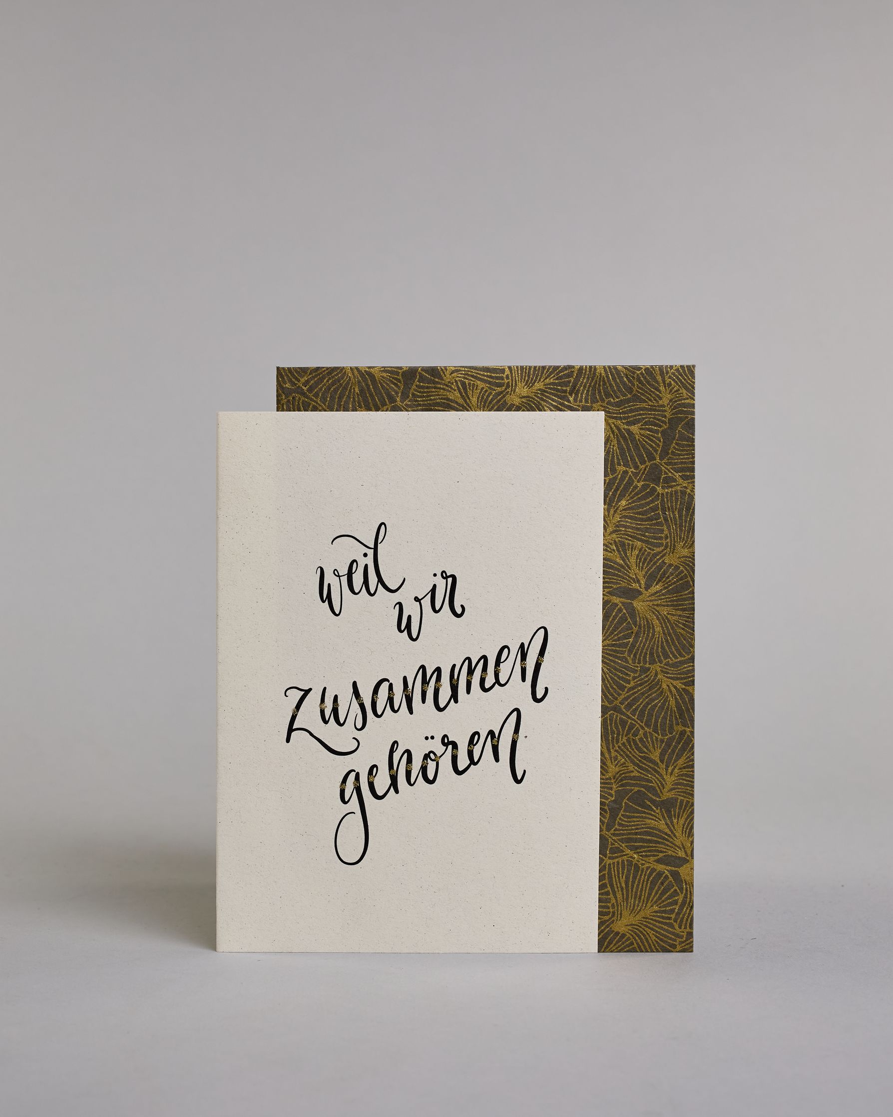 Klappkarte für Liebende mit schwarzer Kalligrafieaufschrift "weil wir zusammen gehören" auf hellem Hintergrund. Der Text ist in vier Zeilen mit jeweils einem Worrt aufgeteilt. Der Text ist in einer geschwungenen Schreibschrift mit punktueller Glitzerverzierung in der Mitte der Buchstaben gestaltet. Dazu ein grün-goldenes handgeschöpftes Kuvert. #2