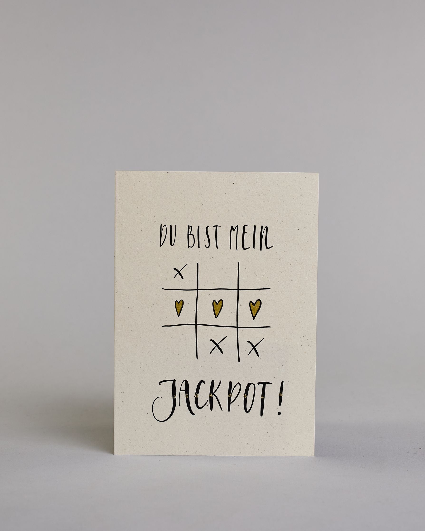 Klappkarte für Liebende mit schwarzer Kalligrafieaufschrift "Du bist mein Jackpot!" mit gezeichnetem Tic-Tac-Toe Herzen statt Kreisen in der mittleren Reihe die das Spiel gewinnen auf hellem Hintergrund. Der Text ist in folgende zwei Zeilen aufgeteilt: "DU BIST MEIN" in verspielter gerader Kalligrafieschrift, darunter das Tic-Tac-Toe Spiel, die Herzen sind gold bemalt, darunter "JACKPOT!" in der gleichen Schrift wie die erste Zeile, nur etwas größer gestaltet und mit punktueller Glitzerverzierung in der Mi…