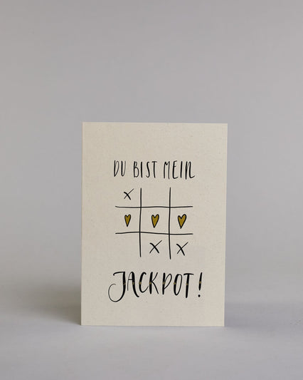 Klappkarte für Liebende mit schwarzer Kalligrafieaufschrift "Du bist mein Jackpot!" mit gezeichnetem Tic-Tac-Toe Herzen statt Kreisen in der mittleren Reihe die das Spiel gewinnen auf hellem Hintergrund. Der Text ist in folgende zwei Zeilen aufgeteilt: "DU BIST MEIN" in verspielter gerader Kalligrafieschrift, darunter das Tic-Tac-Toe Spiel, die Herzen sind gold bemalt, darunter "JACKPOT!" in der gleichen Schrift wie die erste Zeile, nur etwas größer gestaltet und mit punktueller Glitzerverzierung in der Mi…