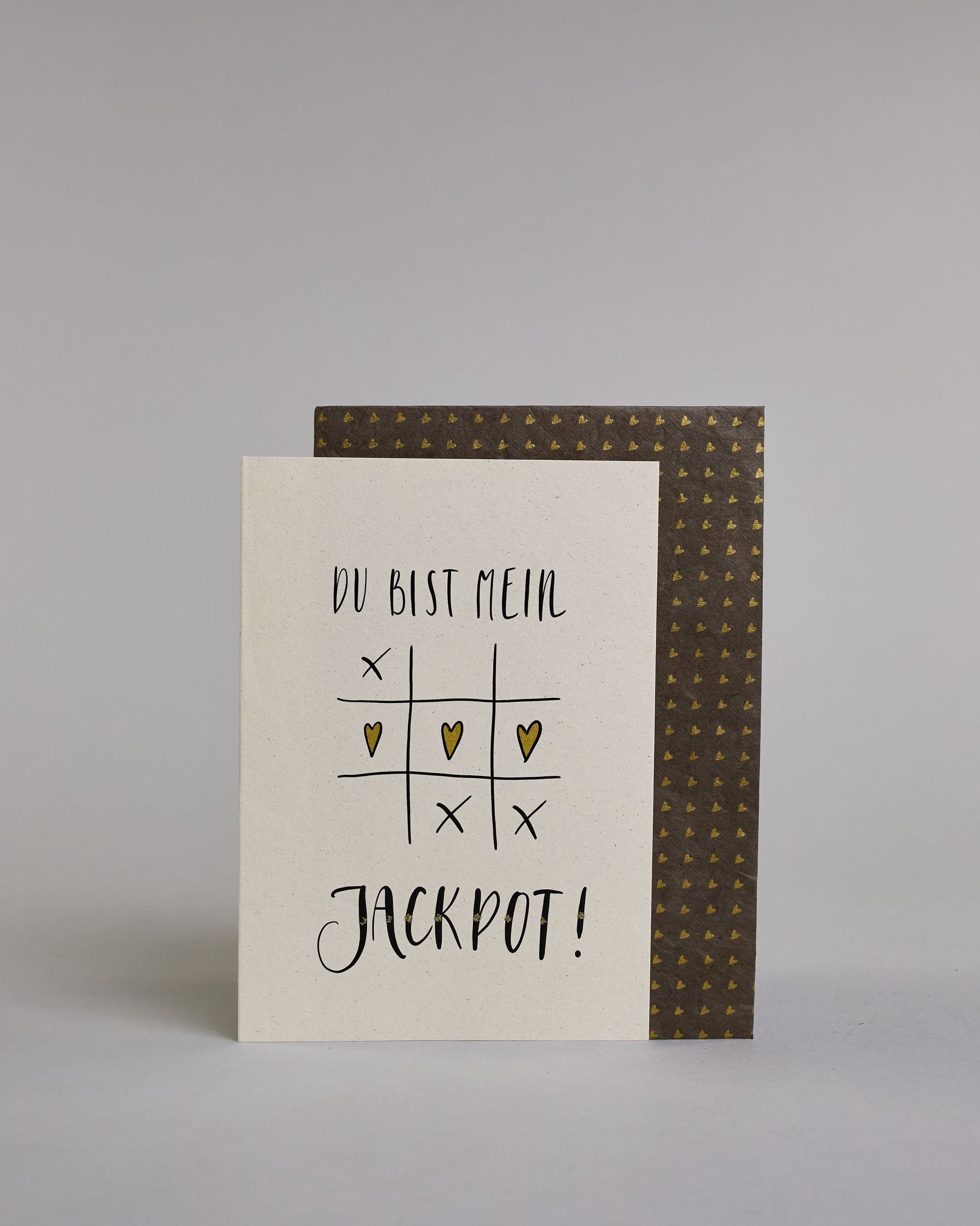 Klappkarte für Liebende mit schwarzer Kalligrafieaufschrift "Du bist mein Jackpot!" mit gezeichnetem Tic-Tac-Toe Herzen statt Kreisen in der mittleren Reihe die das Spiel gewinnen auf hellem Hintergrund. Der Text ist in folgende zwei Zeilen aufgeteilt: "DU BIST MEIN" in verspielter gerader Kalligrafieschrift, darunter das Tic-Tac-Toe Spiel, die Herzen sind gold bemalt, darunter "JACKPOT!" in der gleichen Schrift wie die erste Zeile, nur etwas größer gestaltet und mit punktueller Glitzerverzierung in der Mi…