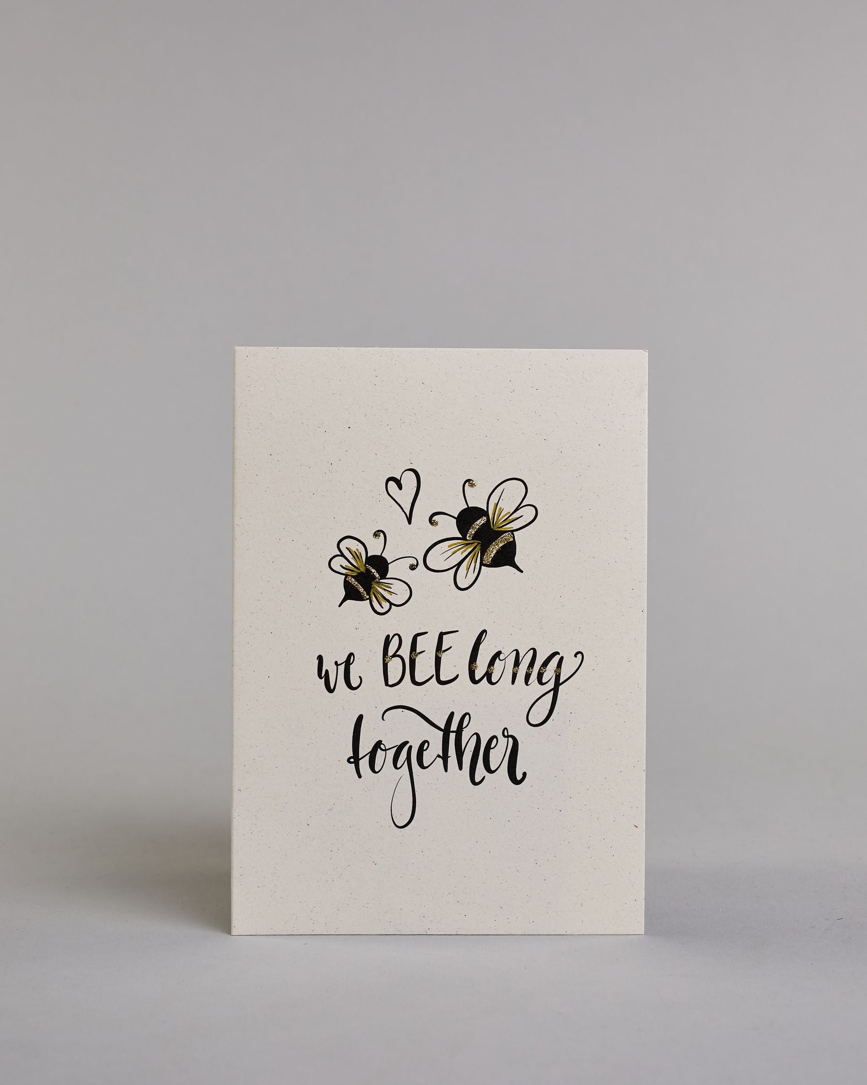 Klappkarte für Liebende in Englisch mit schwarzer Kalligrafieaufschrift "we belong together" mit zwei gezeichneten Bienen und einem Herz über dem Text auf hellem Hintergrund. Der Text ist in folgende zwei Zeilen aufgeteilt: "we BEE long" - das "BEE" ist als einziges Wort in Großbuchstaben gestaltet und wie als das Wort "Biene" und nicht "sein" geschrieben, darunter "together". Der Text ist in verspielter Schreibschrift gestaltet. Dazu ein handgeschöpftes Kuvert. #1