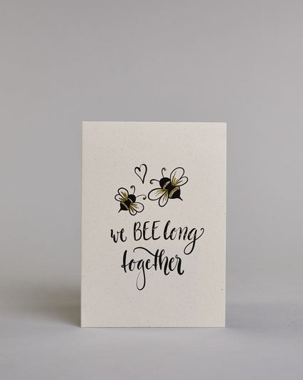 Klappkarte für Liebende in Englisch mit schwarzer Kalligrafieaufschrift "we belong together" mit zwei gezeichneten Bienen und einem Herz über dem Text auf hellem Hintergrund. Der Text ist in folgende zwei Zeilen aufgeteilt: "we BEE long" - das "BEE" ist als einziges Wort in Großbuchstaben gestaltet und wie als das Wort "Biene" und nicht "sein" geschrieben, darunter "together". Der Text ist in verspielter Schreibschrift gestaltet. Dazu ein handgeschöpftes Kuvert. #1