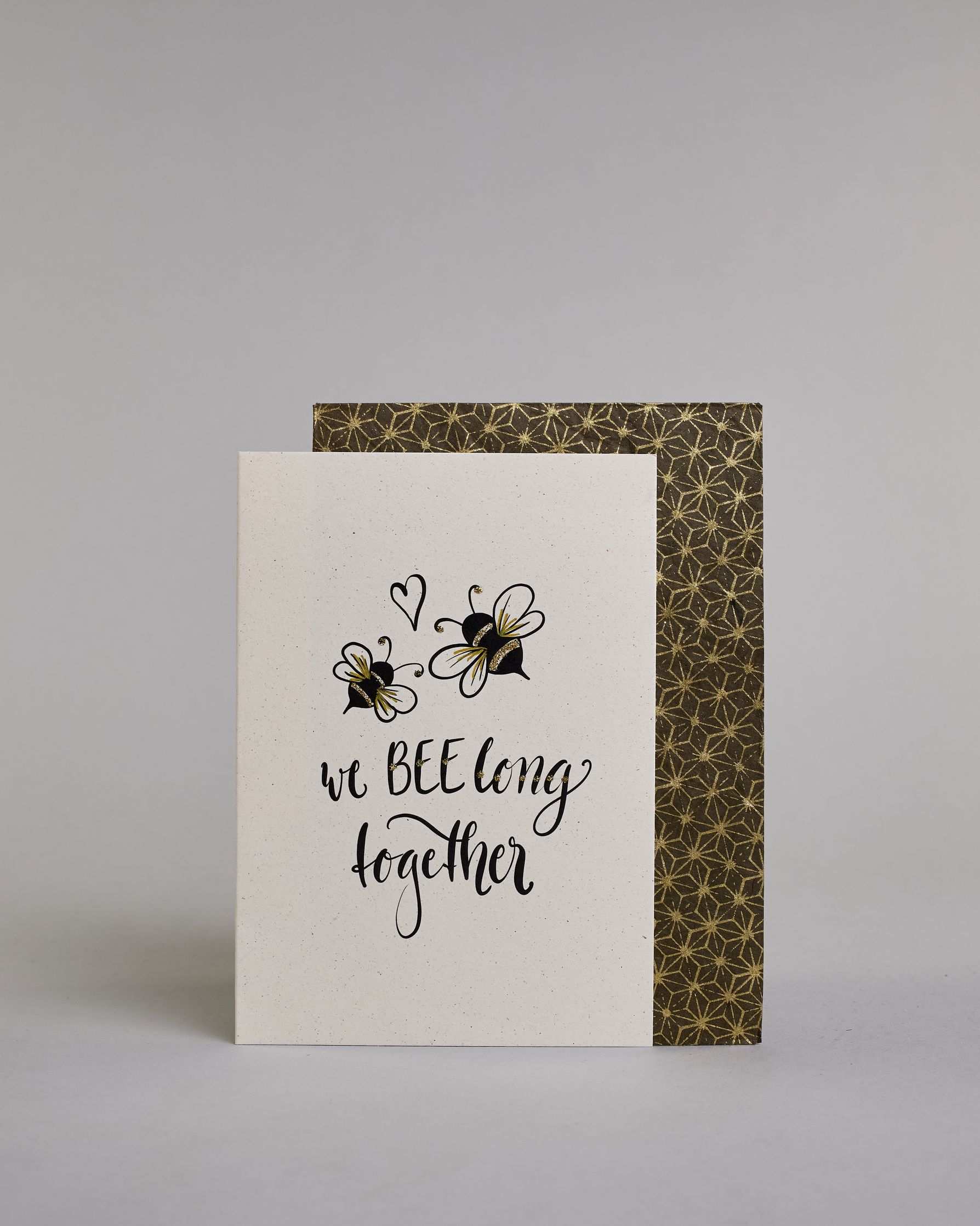 Klappkarte für Liebende in Englisch mit schwarzer Kalligrafieaufschrift "we belong together" mit zwei gezeichneten Bienen und einem Herz über dem Text auf hellem Hintergrund. Der Text ist in folgende zwei Zeilen aufgeteilt: "we BEE long" - das "BEE" ist als einziges Wort in Großbuchstaben gestaltet und wie als das Wort "Biene" und nicht "sein" geschrieben, darunter "together". Der Text ist in verspielter Schreibschrift gestaltet. Dazu ein handgeschöpftes Kuvert. #2