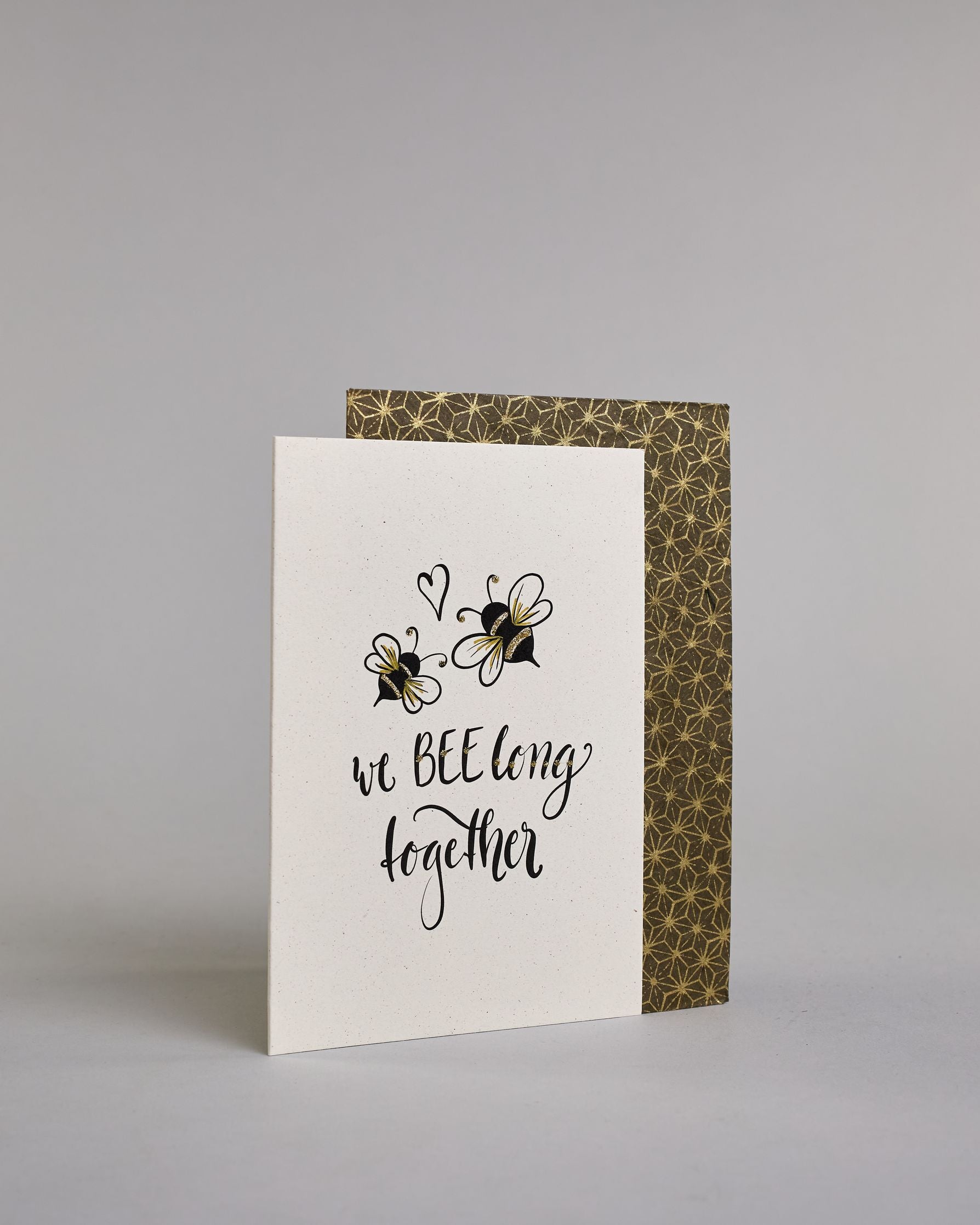 Klappkarte für Liebende in Englisch mit schwarzer Kalligrafieaufschrift "we belong together" mit zwei gezeichneten Bienen und einem Herz über dem Text auf hellem Hintergrund. Der Text ist in folgende zwei Zeilen aufgeteilt: "we BEE long" - das "BEE" ist als einziges Wort in Großbuchstaben gestaltet und wie als das Wort "Biene" und nicht "sein" geschrieben, darunter "together". Der Text ist in verspielter Schreibschrift gestaltet. Dazu ein handgeschöpftes Kuvert. #3