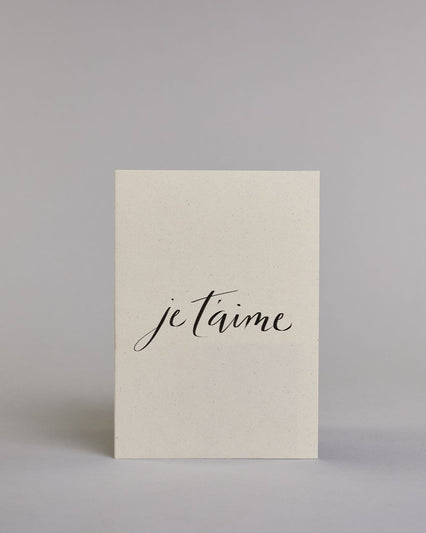 Klappkarte für Liebende auf Französisch mit schwarzer Kalligrafieaufschrift "je t'aime" auf hellem Hintergrund. Der Text ist in einer Zeile und in einer Schreibschrift gestaltet. Dazu ein rot-weißes handgeschöpftes Kuvert. #1