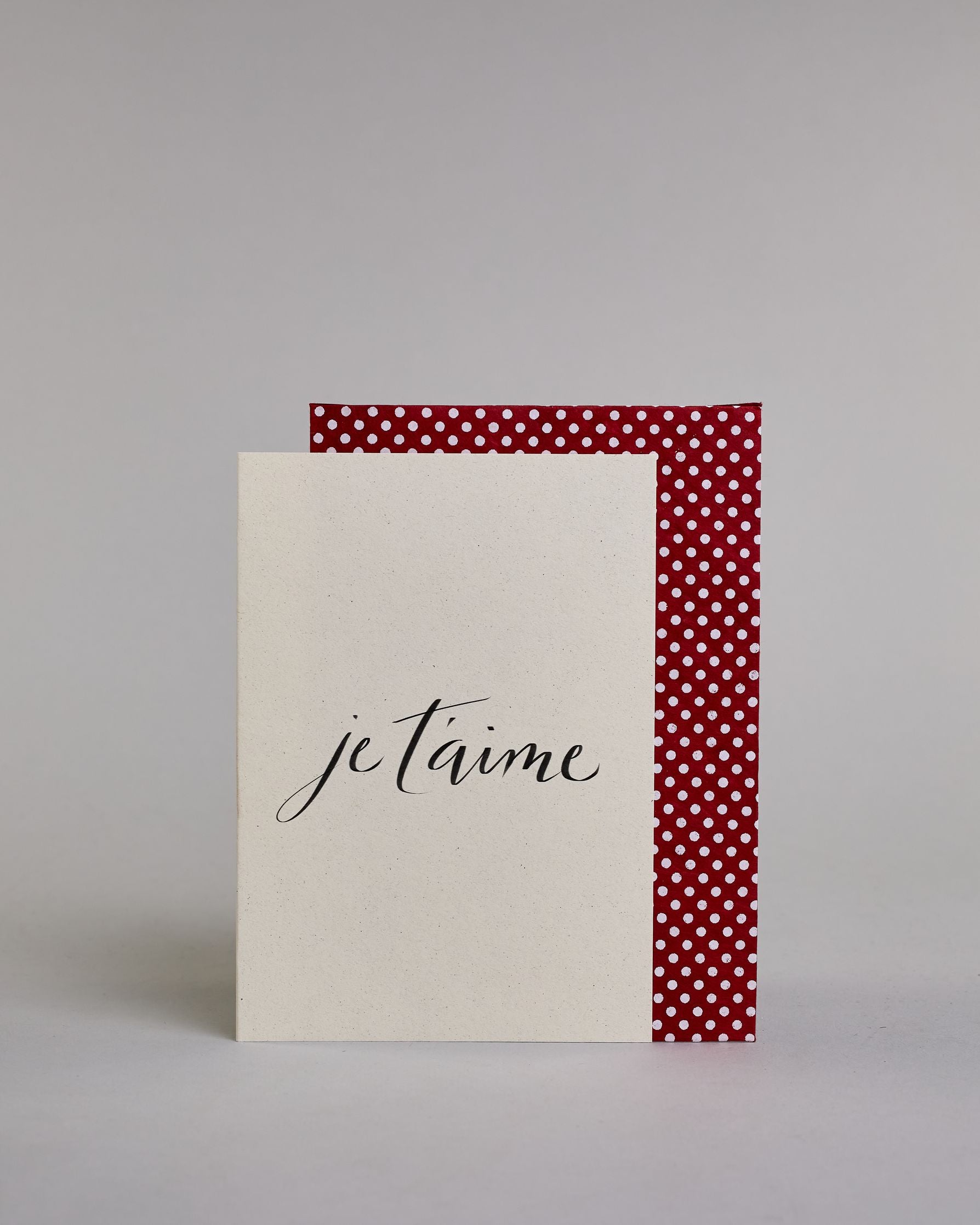 Klappkarte für Liebende auf Französisch mit schwarzer Kalligrafieaufschrift "je t'aime" auf hellem Hintergrund. Der Text ist in einer Zeile und in einer Schreibschrift gestaltet. Dazu ein rot-weißes handgeschöpftes Kuvert. #2