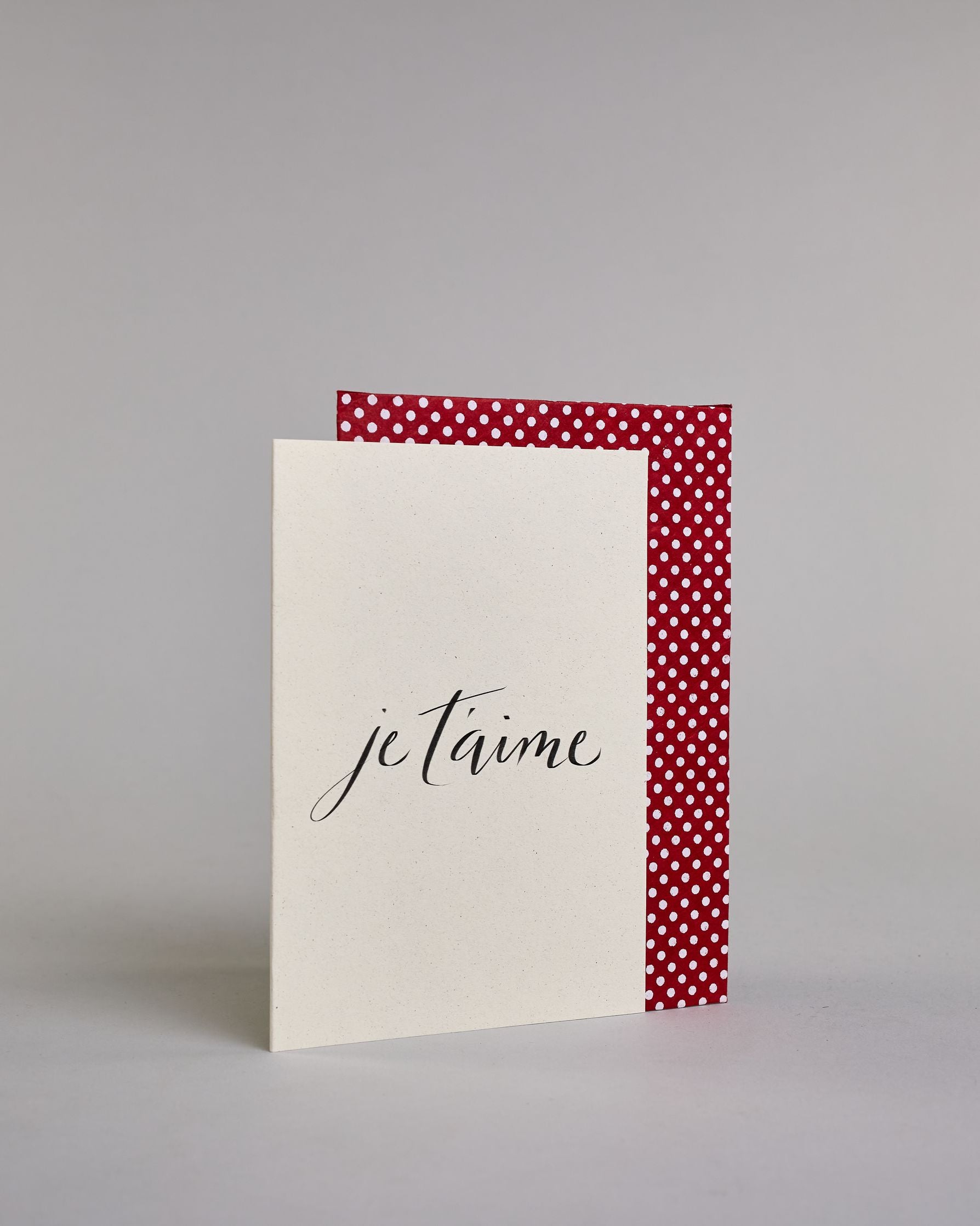 Klappkarte für Liebende auf Französisch mit schwarzer Kalligrafieaufschrift "je t'aime" auf hellem Hintergrund. Der Text ist in einer Zeile und in einer Schreibschrift gestaltet. Dazu ein rot-weißes handgeschöpftes Kuvert. #3
