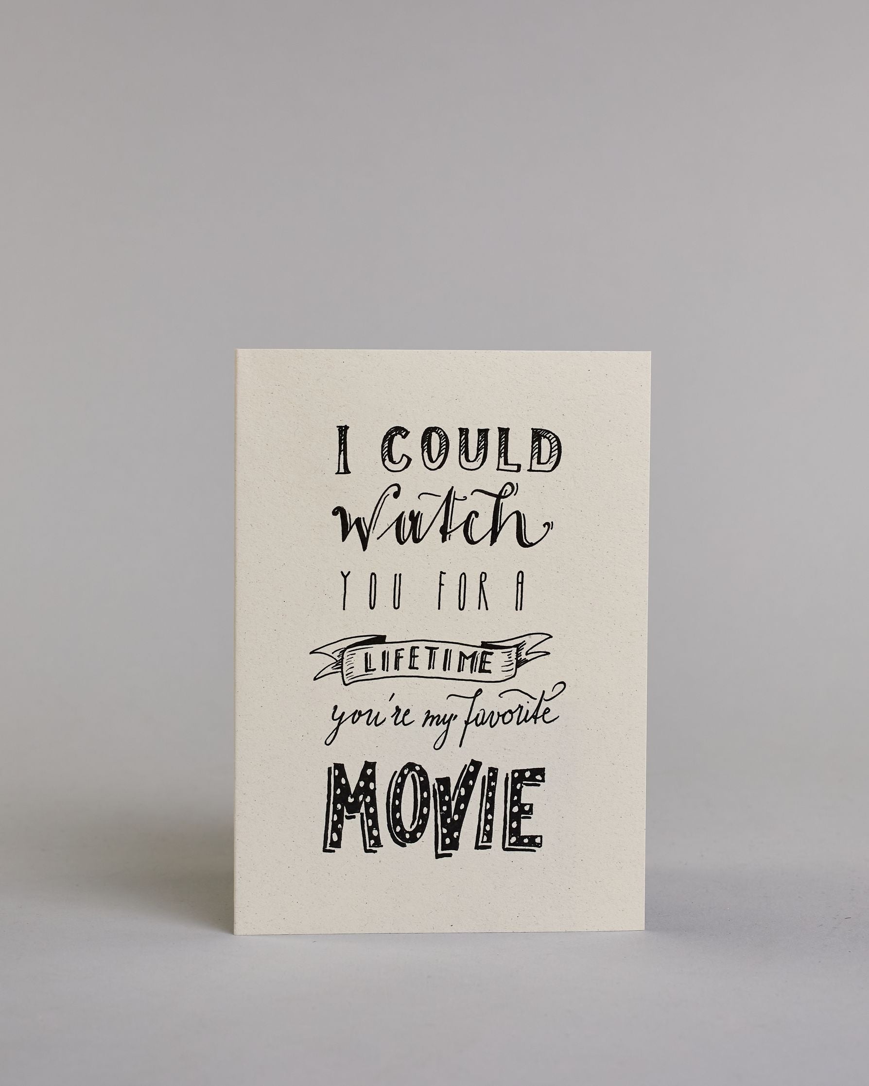 Klappkarte für Liebende auf Englisch mit schwarzer Kalligrafieaufschrift "I could watch you for a lifetime you're my favorite movie" auf hellem Hintergrund. Der Text ist in folgende sechs Zeilen aufgeteilt: "I COULD" in dicker gerader Kalligrafieschrift, darunter "watch" in geschwungener Schreibschrift, darunter "YOU FOR A" in dünner gerader Kalligrafieschrift, darunter "LIFETIME" eingefasst in einem Banner, darunter "you're my favorite" in kleiner Schreibschrift, darunter "MOVIE" in dicken großen Blockbuc…