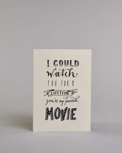 Klappkarte für Liebende auf Englisch mit schwarzer Kalligrafieaufschrift "I could watch you for a lifetime you're my favorite movie" auf hellem Hintergrund. Der Text ist in folgende sechs Zeilen aufgeteilt: "I COULD" in dicker gerader Kalligrafieschrift, darunter "watch" in geschwungener Schreibschrift, darunter "YOU FOR A" in dünner gerader Kalligrafieschrift, darunter "LIFETIME" eingefasst in einem Banner, darunter "you're my favorite" in kleiner Schreibschrift, darunter "MOVIE" in dicken großen Blockbuc…