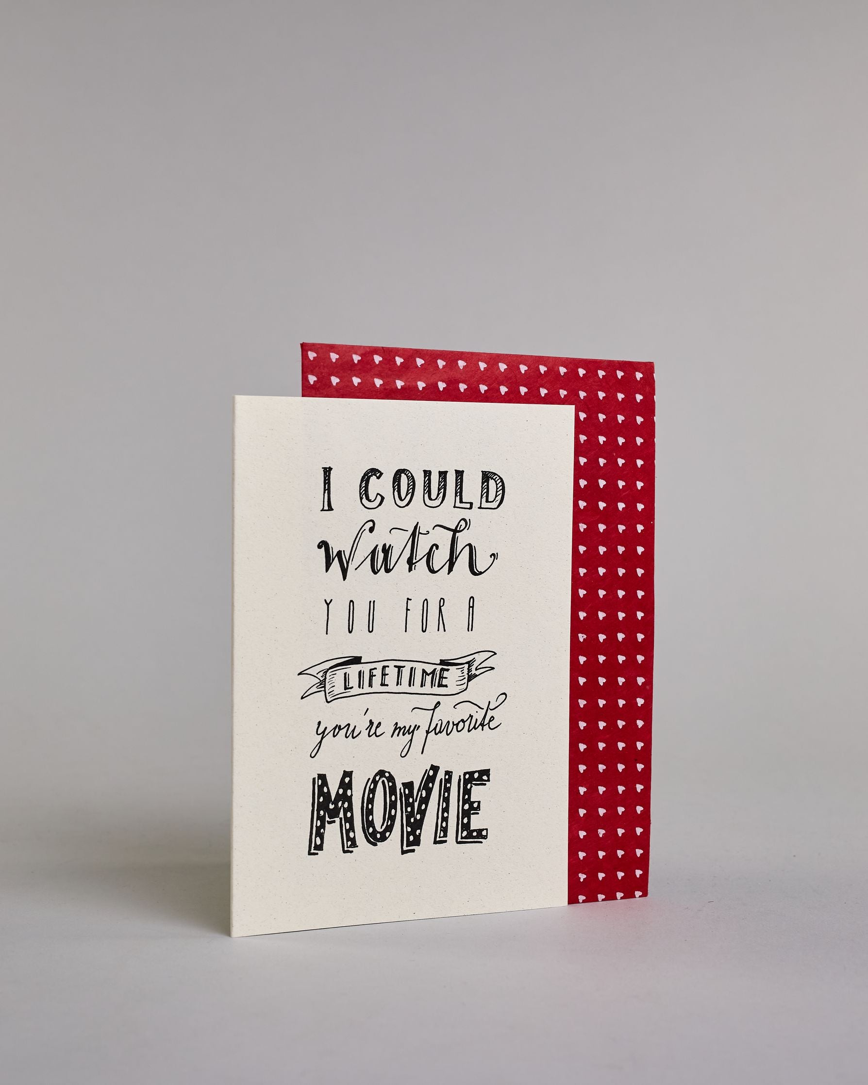 Klappkarte für Liebende auf Englisch mit schwarzer Kalligrafieaufschrift "I could watch you for a lifetime you're my favorite movie" auf hellem Hintergrund. Der Text ist in folgende sechs Zeilen aufgeteilt: "I COULD" in dicker gerader Kalligrafieschrift, darunter "watch" in geschwungener Schreibschrift, darunter "YOU FOR A" in dünner gerader Kalligrafieschrift, darunter "LIFETIME" eingefasst in einem Banner, darunter "you're my favorite" in kleiner Schreibschrift, darunter "MOVIE" in dicken großen Blockbuc…