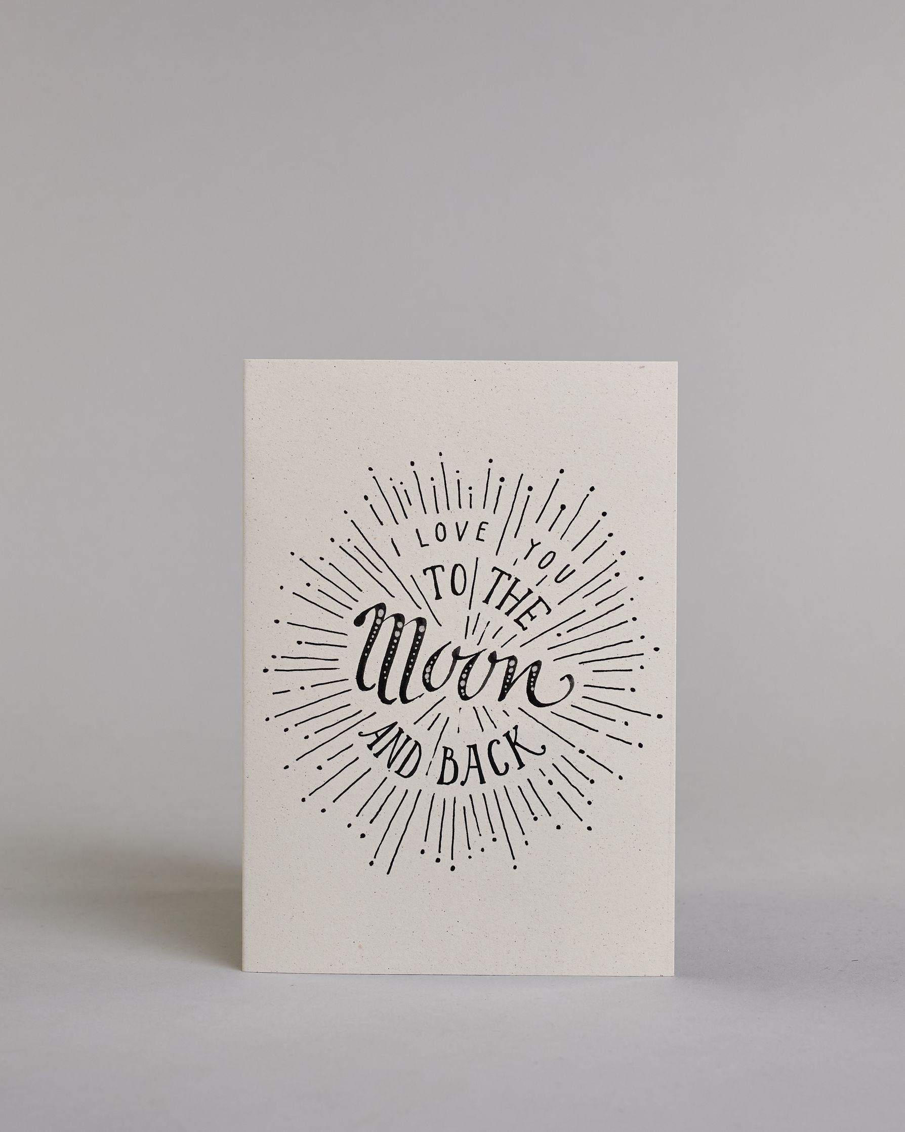 Klappkarte für Liebende auf Englisch mit schwarzer Kalligrafieaufschrift "I love you to the moon and back" auf hellem Hintergrund. Der Text ist in folgende vier Zeilen aufgeteilt: "I lOVE YOU" in dünner gerader Kalligrafieschrift, darunter "TO THE" in größerer gerader Kalligrafieschrift, darunter "Moon" in kunstvoller Schreibschrift, darunter "AND BACK" and verspielter gerader Kalligrafieschrift. Der Text ist umgeben von einzelnen Linien und Punkten die im Gesamtbild einen Kreis, ähnlich einer Explosion, e…