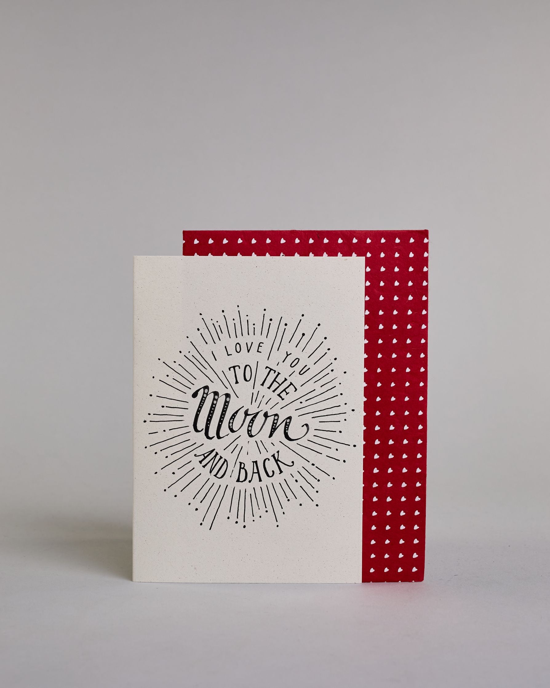 Klappkarte für Liebende auf Englisch mit schwarzer Kalligrafieaufschrift "I love you to the moon and back" auf hellem Hintergrund. Der Text ist in folgende vier Zeilen aufgeteilt: "I lOVE YOU" in dünner gerader Kalligrafieschrift, darunter "TO THE" in größerer gerader Kalligrafieschrift, darunter "Moon" in kunstvoller Schreibschrift, darunter "AND BACK" and verspielter gerader Kalligrafieschrift. Der Text ist umgeben von einzelnen Linien und Punkten die im Gesamtbild einen Kreis, ähnlich einer Explosion, e…