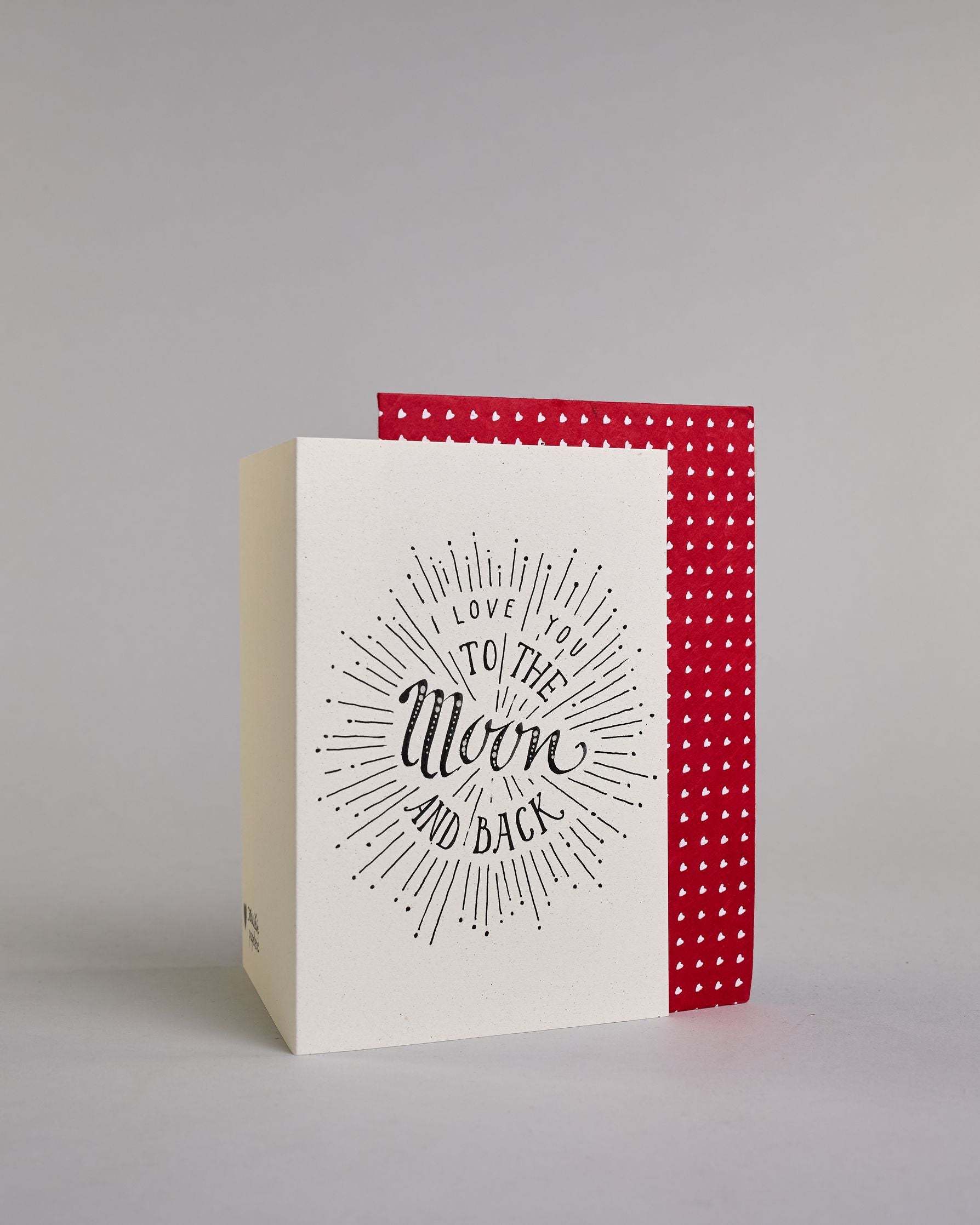 Klappkarte für Liebende auf Englisch mit schwarzer Kalligrafieaufschrift "I love you to the moon and back" auf hellem Hintergrund. Der Text ist in folgende vier Zeilen aufgeteilt: "I lOVE YOU" in dünner gerader Kalligrafieschrift, darunter "TO THE" in größerer gerader Kalligrafieschrift, darunter "Moon" in kunstvoller Schreibschrift, darunter "AND BACK" and verspielter gerader Kalligrafieschrift. Der Text ist umgeben von einzelnen Linien und Punkten die im Gesamtbild einen Kreis, ähnlich einer Explosion, e…