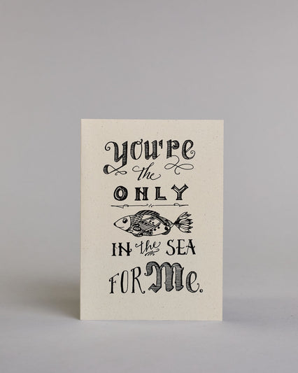 Klappkarte für Liebende auf Englisch mit schwarzer Kalligrafieaufschrift "you're the only fish in the sea for me" mit einem gezeichneten Fisch auf hellem Hintergrund. Der Text ist in folgende fünf Zeilen aufgeteilt: "you're" in geschwungener kunstvoller Kalligrafieschrift, darunter "the" in dünner Schreibschrift zwischen zwei Schnörkeln, darunter "ONLY" in kunstvoller Kalligrafieschrift, darunter eine horizontale Linie, darunter ein kunstvoll gestalteter gezeichneter Fisch, darunter "IN the SEA" in verspie…