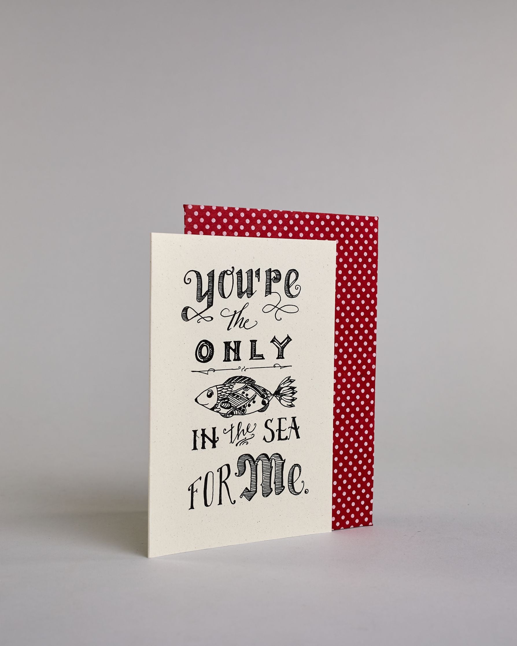 Klappkarte für Liebende auf Englisch mit schwarzer Kalligrafieaufschrift "you're the only fish in the sea for me" mit einem gezeichneten Fisch auf hellem Hintergrund. Der Text ist in folgende fünf Zeilen aufgeteilt: "you're" in geschwungener kunstvoller Kalligrafieschrift, darunter "the" in dünner Schreibschrift zwischen zwei Schnörkeln, darunter "ONLY" in kunstvoller Kalligrafieschrift, darunter eine horizontale Linie, darunter ein kunstvoll gestalteter gezeichneter Fisch, darunter "IN the SEA" in verspie…