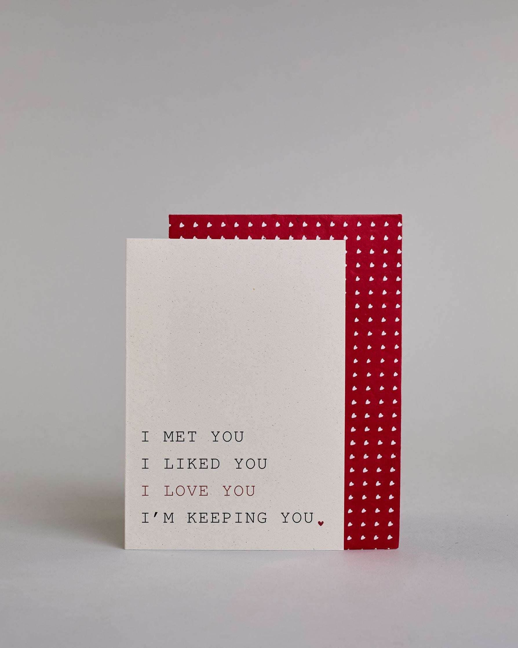 Klappkarte für Liebende auf Englisch mit schwarzer und roter Kalligrafieaufschrift "I met you I liked you I love you I'm keeping you." auf hellem Hintergrund. Der Text ist in folgende vier Zeilen aufgeteilt: "I MET YOU" in schwarzer, darunter "I LIKED YOU" in schwarzer, darunter "I LOVE YOU" in roter, darunter "I'M KEEPING YOU." in schwarzer Schrift, der Punkt hinter dem "YOU" ist ein kleines rotes Herz. Alle Zeile sind in einer Kalligrafieschrit gestaltet die an die tyische Schreibmaschinenschrift erinner…