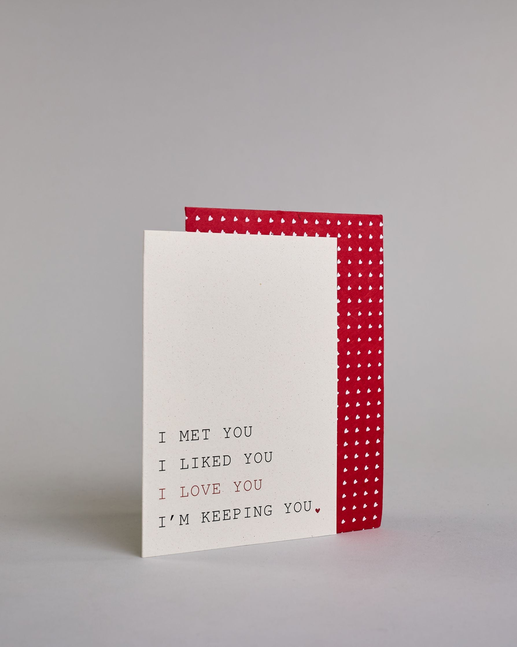 Klappkarte für Liebende auf Englisch mit schwarzer und roter Kalligrafieaufschrift "I met you I liked you I love you I'm keeping you." auf hellem Hintergrund. Der Text ist in folgende vier Zeilen aufgeteilt: "I MET YOU" in schwarzer, darunter "I LIKED YOU" in schwarzer, darunter "I LOVE YOU" in roter, darunter "I'M KEEPING YOU." in schwarzer Schrift, der Punkt hinter dem "YOU" ist ein kleines rotes Herz. Alle Zeile sind in einer Kalligrafieschrit gestaltet die an die tyische Schreibmaschinenschrift erinner…