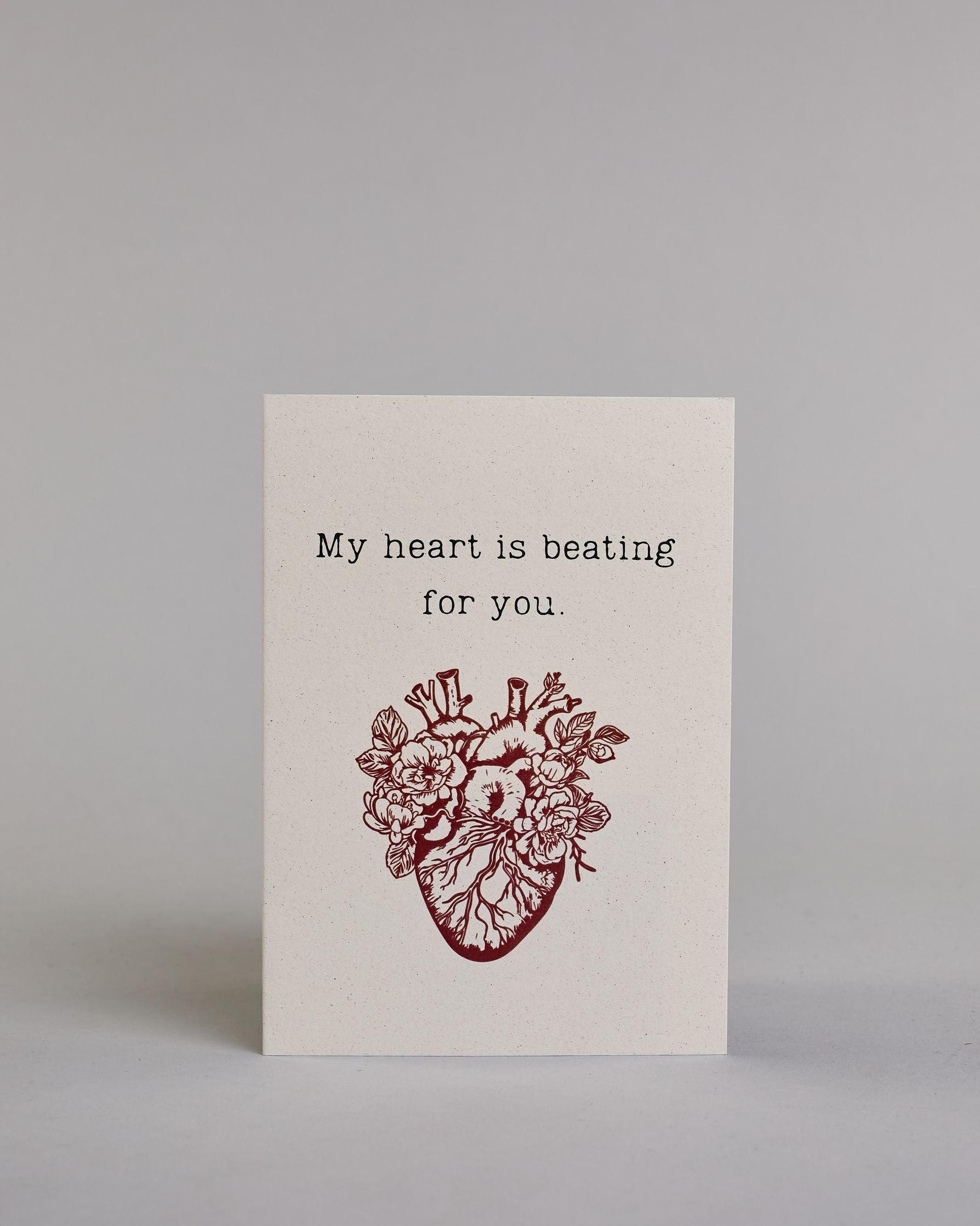 Klappkarte für Liebende auf Englisch mit schwarzer und Kalligrafieaufschrift "My heart is beating for you." mit einem rotem gezeichnetem Herz das einem echten Herz nachempfunden ist, allerdings ist es mit Blüten und Blättern verschönert auf hellem Hintergrund. Der Text ist in folgende zwei Zeilen aufgeteilt: "My heart is beating", darunter "for you." - beide Zeile in typischer Schreibmaschinenschrift, darunter das rote Herz. Dazu ein rot-weißes handgeschöpftes Kuvert. #1