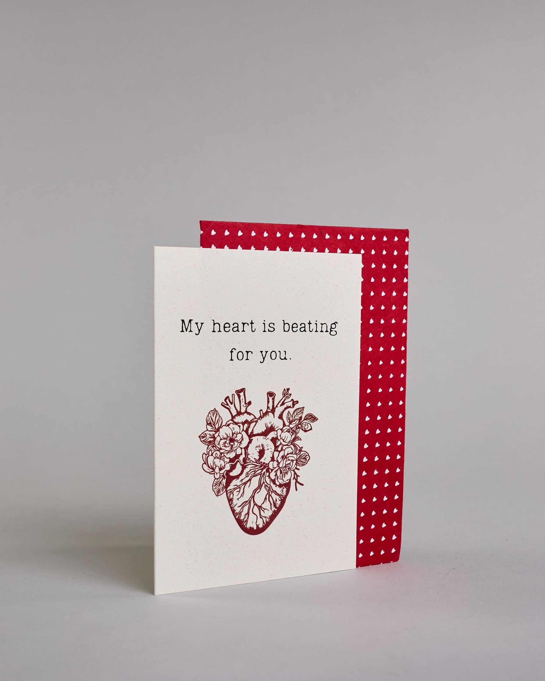 Klappkarte für Liebende auf Englisch mit schwarzer und Kalligrafieaufschrift "My heart is beating for you." mit einem rotem gezeichnetem Herz das einem echten Herz nachempfunden ist, allerdings ist es mit Blüten und Blättern verschönert auf hellem Hintergrund. Der Text ist in folgende zwei Zeilen aufgeteilt: "My heart is beating", darunter "for you." - beide Zeile in typischer Schreibmaschinenschrift, darunter das rote Herz. Dazu ein rot-weißes handgeschöpftes Kuvert. #3