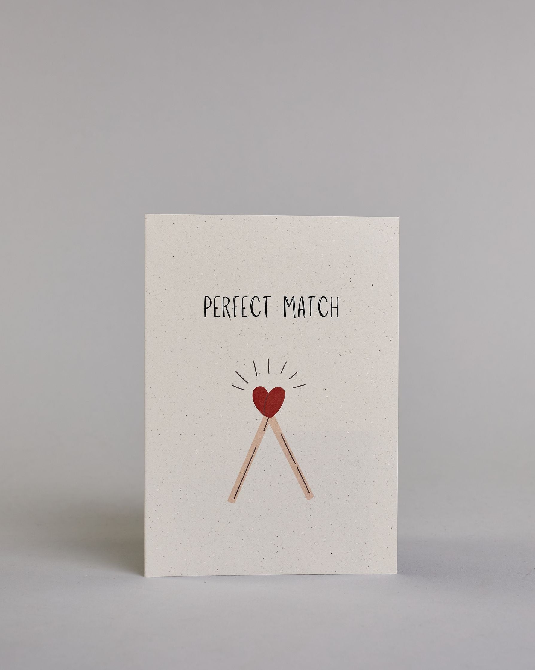Klappkarte für Liebende auf Englisch mit schwarzer und Kalligrafieaufschrift "Perfect Match" mit zwei gezeichneten Streichhölzern, dessen Köpfe ein Herz ergeben auf hellem Hintergrund. Der Text ist in einer Zeile zusammengefasst und in einer gerade dünnen Kalligrafieschrift gestaltet. Darunter sind die Streichhölzer dessen Stil ein helles braun ist und dessen Köpfe, die das Herz ergeben rot sind, über den Köpfen sind einzelne Linien die wie ein Kranz von der Zeichnung wegführen. Dazu ein rot-weißes handges…