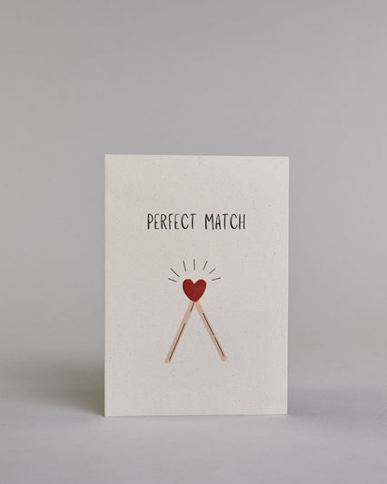Klappkarte für Liebende auf Englisch mit schwarzer und Kalligrafieaufschrift "Perfect Match" mit zwei gezeichneten Streichhölzern, dessen Köpfe ein Herz ergeben auf hellem Hintergrund. Der Text ist in einer Zeile zusammengefasst und in einer gerade dünnen Kalligrafieschrift gestaltet. Darunter sind die Streichhölzer dessen Stil ein helles braun ist und dessen Köpfe, die das Herz ergeben rot sind, über den Köpfen sind einzelne Linien die wie ein Kranz von der Zeichnung wegführen. Dazu ein rot-weißes handges…