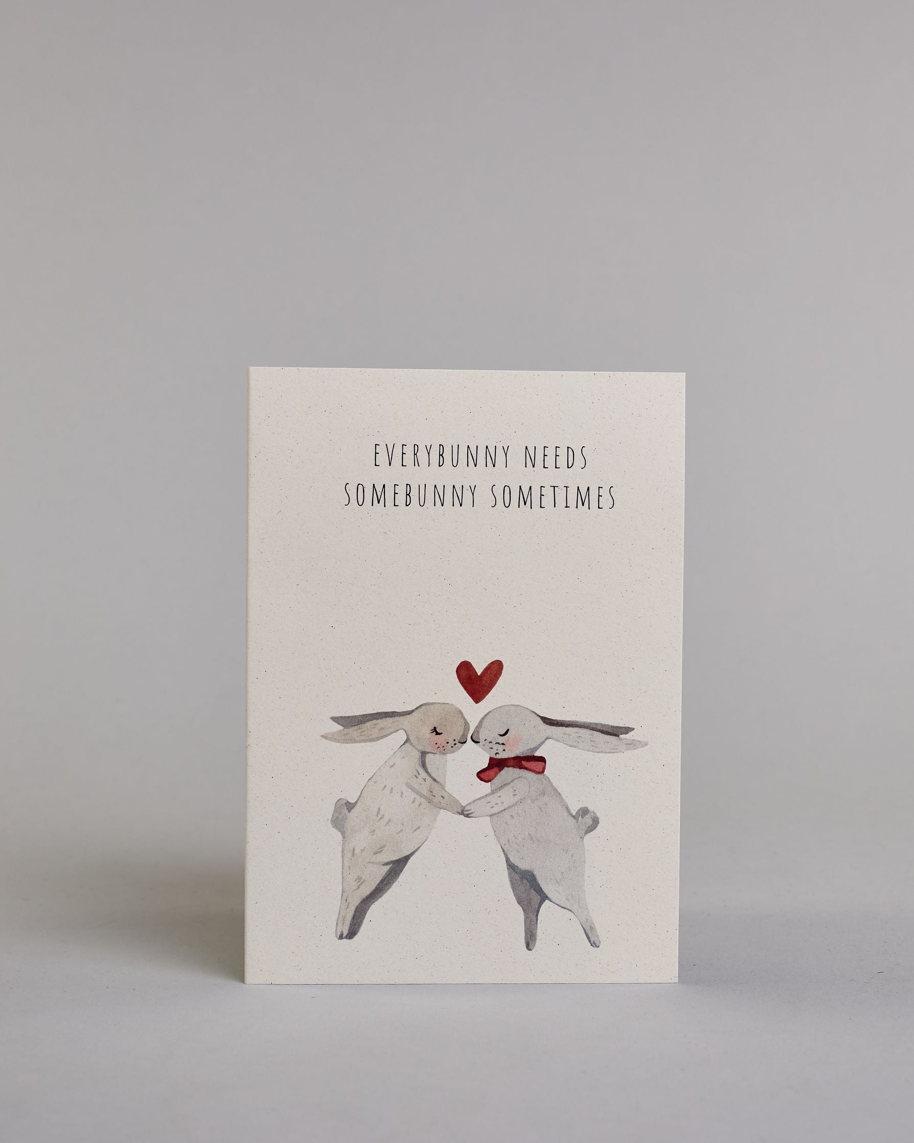 Klappkarte für Liebende auf Englisch mit schwarzer Kalligrafieaufschrift "Everybunny needs somebunny sometimes", darunter sind zwei gezeichnete Hasen in einem hellen beige und hellem grau die ihre Nasen aufeinander zuführen auf hellem Hintergrund. Der rechte Hase hat eine rote Masche um den Hals und über ihren Köpfen ist ein rotes Herz. Die Kalligrafieschrift ist dünn und gerade und teilt sich in zwei Zeilen mit jeweils immer zwei Worten auf. Die Schrift ist im oberen linken Teil der Karte, während die Has…