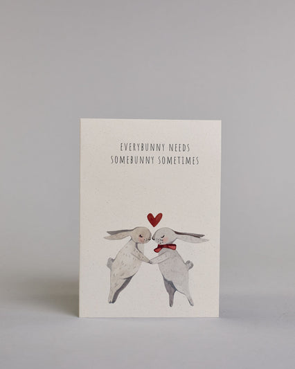 Klappkarte für Liebende auf Englisch mit schwarzer Kalligrafieaufschrift "Everybunny needs somebunny sometimes", darunter sind zwei gezeichnete Hasen in einem hellen beige und hellem grau die ihre Nasen aufeinander zuführen auf hellem Hintergrund. Der rechte Hase hat eine rote Masche um den Hals und über ihren Köpfen ist ein rotes Herz. Die Kalligrafieschrift ist dünn und gerade und teilt sich in zwei Zeilen mit jeweils immer zwei Worten auf. Die Schrift ist im oberen linken Teil der Karte, während die Has…
