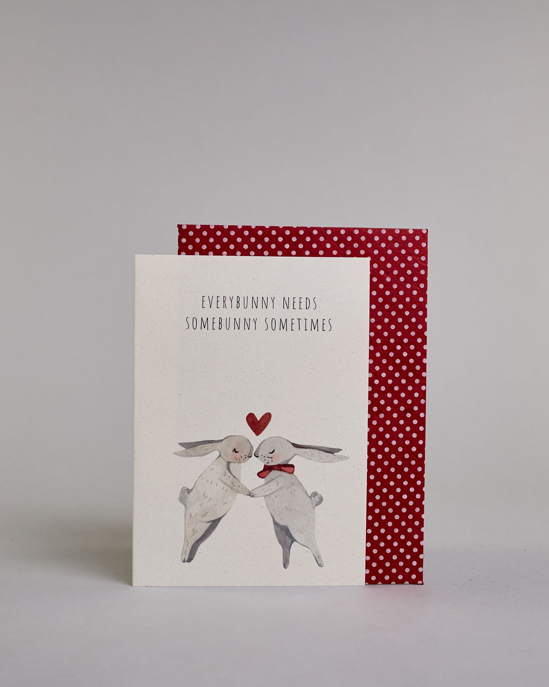 Klappkarte für Liebende auf Englisch mit schwarzer Kalligrafieaufschrift "Everybunny needs somebunny sometimes", darunter sind zwei gezeichnete Hasen in einem hellen beige und hellem grau die ihre Nasen aufeinander zuführen auf hellem Hintergrund. Der rechte Hase hat eine rote Masche um den Hals und über ihren Köpfen ist ein rotes Herz. Die Kalligrafieschrift ist dünn und gerade und teilt sich in zwei Zeilen mit jeweils immer zwei Worten auf. Die Schrift ist im oberen linken Teil der Karte, während die Has…