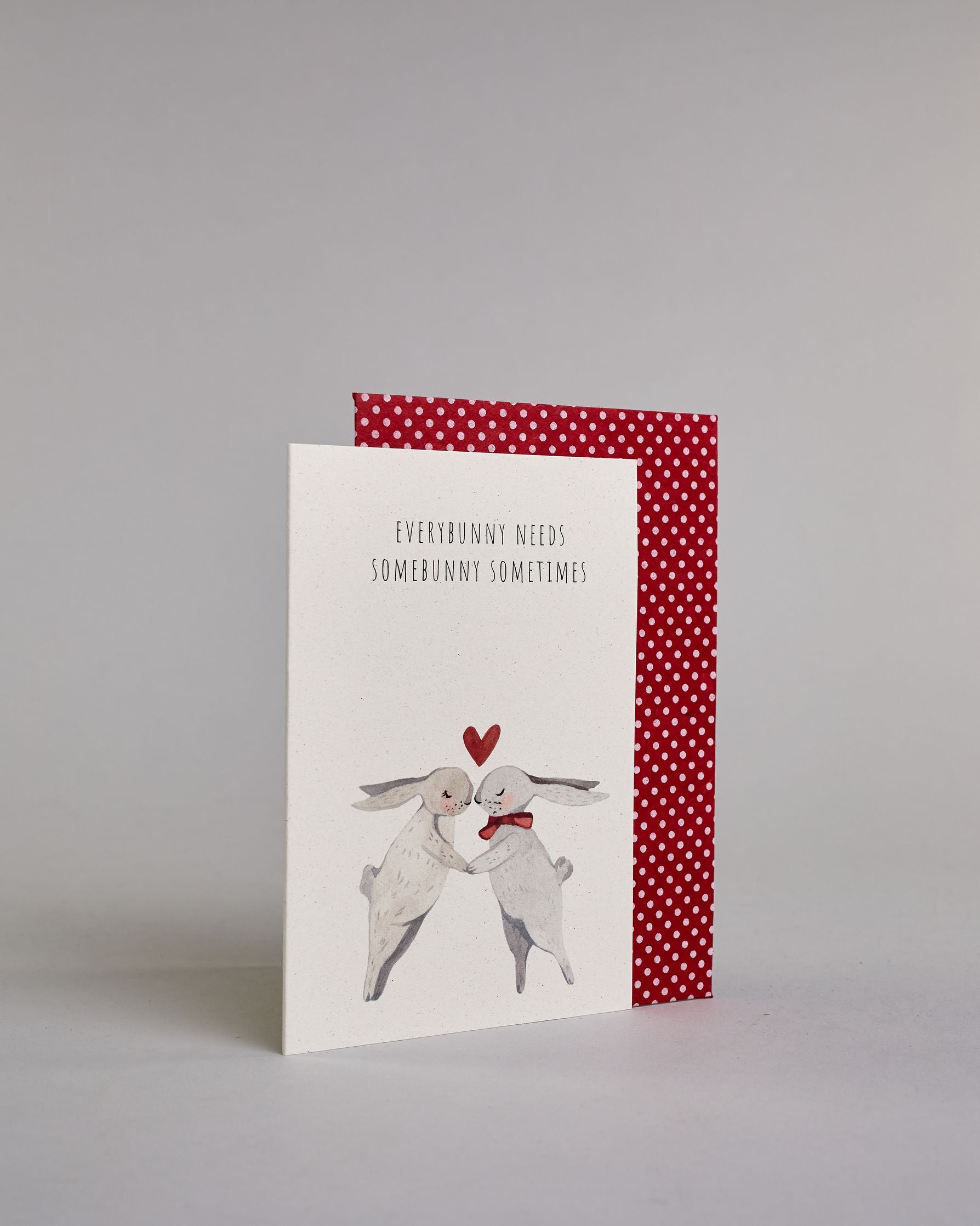 Klappkarte für Liebende auf Englisch mit schwarzer Kalligrafieaufschrift "Everybunny needs somebunny sometimes", darunter sind zwei gezeichnete Hasen in einem hellen beige und hellem grau die ihre Nasen aufeinander zuführen auf hellem Hintergrund. Der rechte Hase hat eine rote Masche um den Hals und über ihren Köpfen ist ein rotes Herz. Die Kalligrafieschrift ist dünn und gerade und teilt sich in zwei Zeilen mit jeweils immer zwei Worten auf. Die Schrift ist im oberen linken Teil der Karte, während die Has…