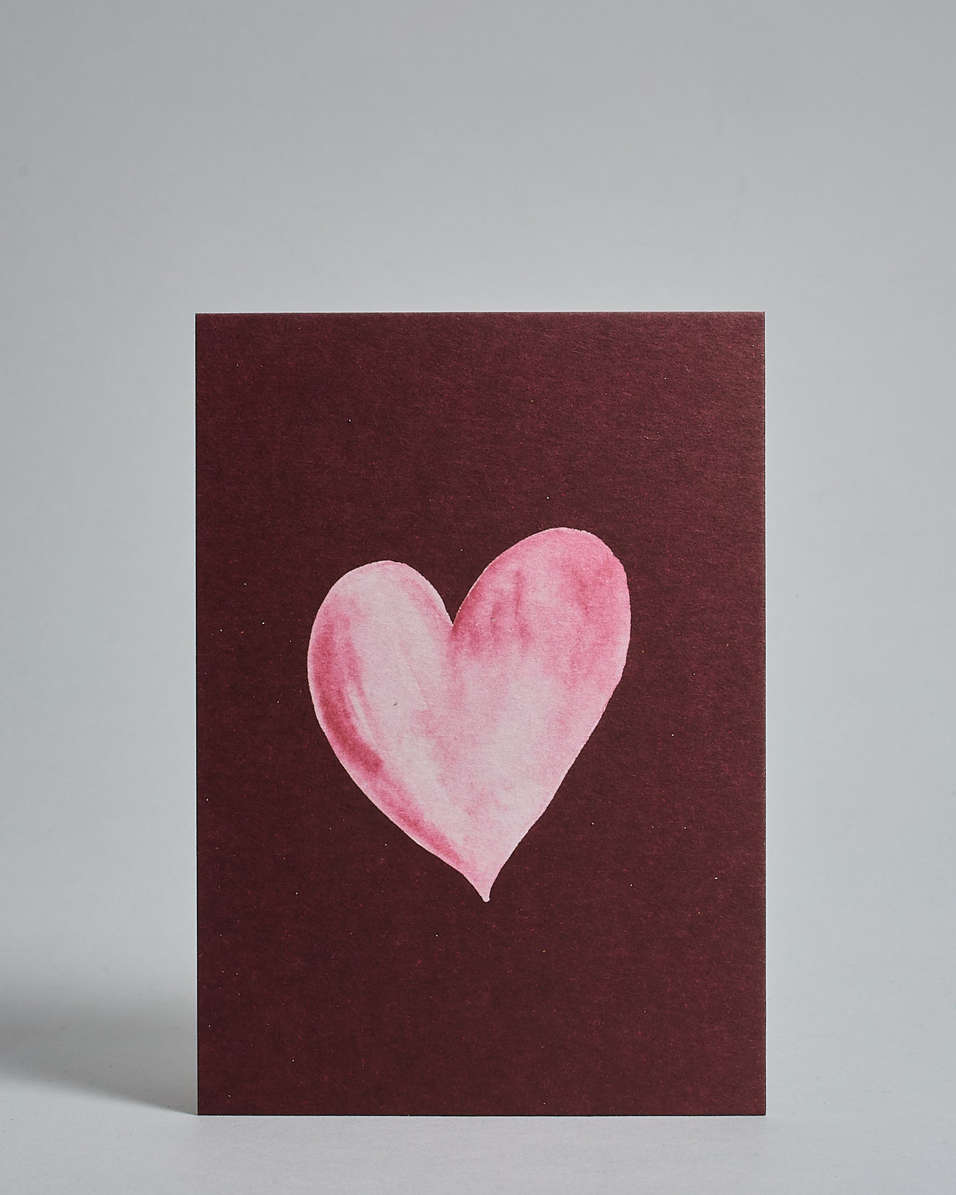 Die Vorderseite der Postkarte im Hochformat vor hellgrauem Hintergrund. Die Postkarte zeigt ein rosa-farbenes, gezeichnet aussehendes Herz auf bordeaux-farbenem Hintergrund, gedruckt auf Holzschliffpappe.