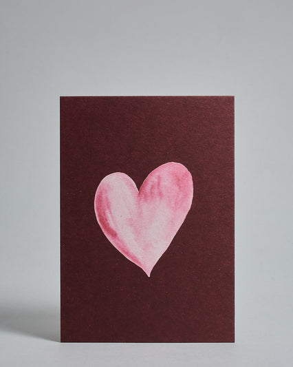 Die Vorderseite der Postkarte im Hochformat vor hellgrauem Hintergrund. Die Postkarte zeigt ein rosa-farbenes, gezeichnet aussehendes Herz auf bordeaux-farbenem Hintergrund, gedruckt auf Holzschliffpappe.