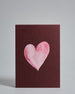 Die Vorderseite der Postkarte im Hochformat vor hellgrauem Hintergrund. Die Postkarte zeigt ein rosa-farbenes, gezeichnet aussehendes Herz auf bordeaux-farbenem Hintergrund, gedruckt auf Holzschliffpappe.