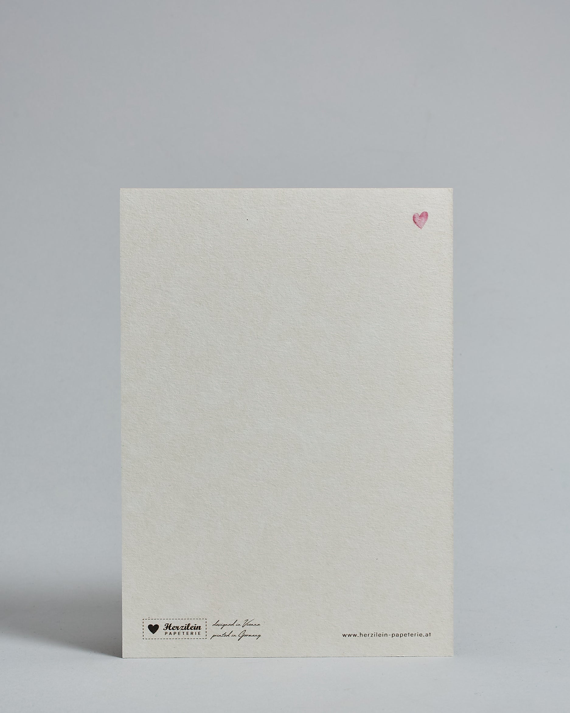 Die Rückseite der Postkarte im Hochformat vor hellgrauem Hintergrund. Rechts oben ist in kleiner Ausführung das gleiche rosa Herz wie auf der Vorderseite zu sehen. 
Unten sind klein das Logo Herzilein-Papeterie, der Text: "designed in VIenna, printed in Germany" und "www.herzilein-papeterie.at" zu sehen. 
Der Großteil ist unbedruckt die Struktur der hochwertigen Holzschliffpappe ist gut zu sehen. Hier ist viel Platz zum Schreiben.