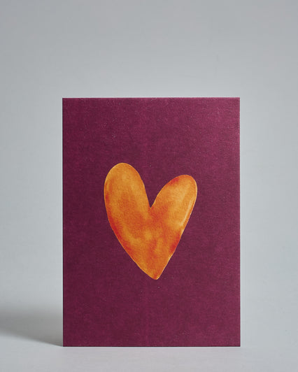 Die Vorderseite der Postkarte im Hochformat vor hellgrauem Hintergrund. Die Postkarte zeigt ein orangene, gezeichnet aussehendes Herz auf purpur-farbenem Hintergrund, gedruckt auf Holzschliffpappe.